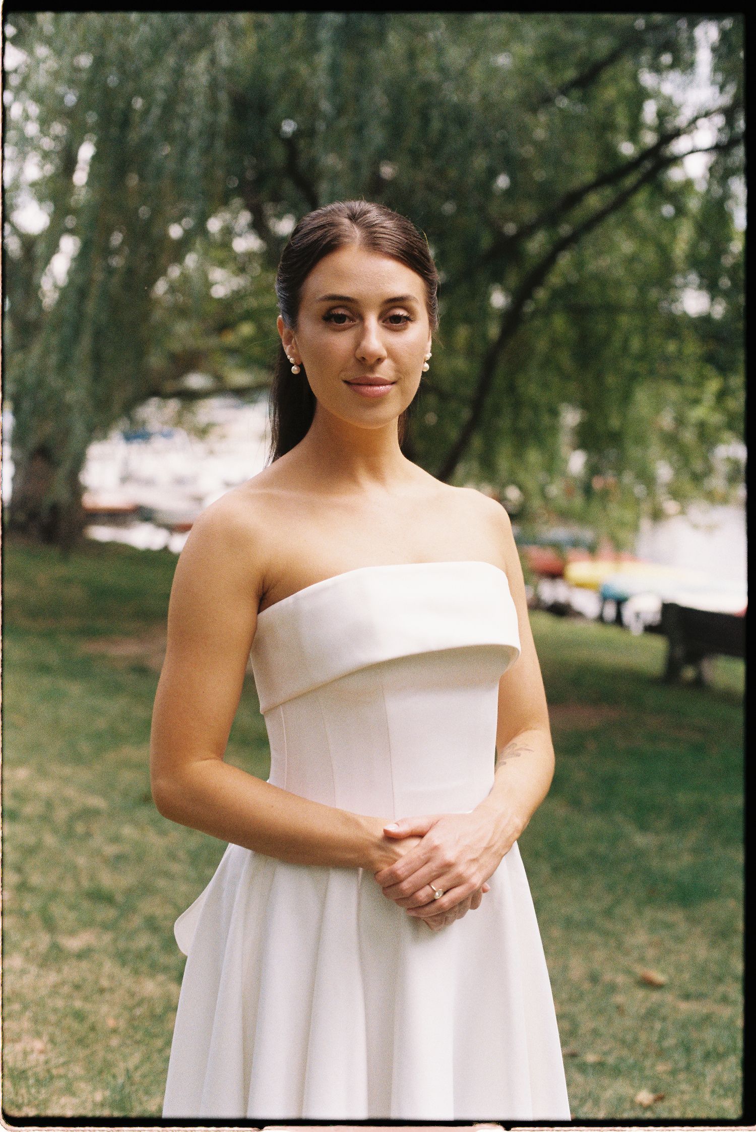 A bride in a white strapless gown poses outdoors in a park with lush green willow trees in the background.