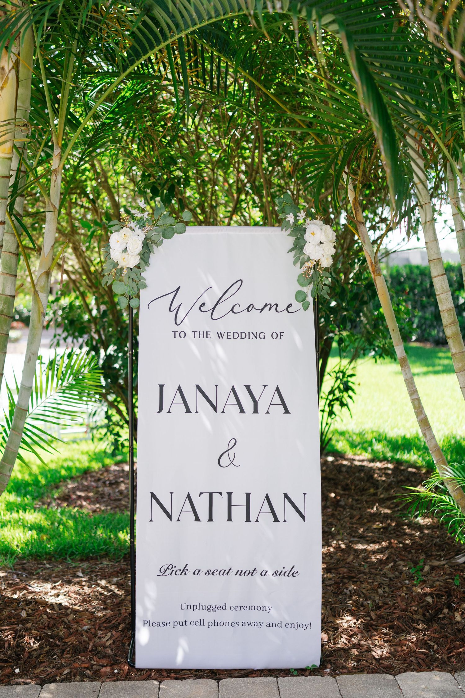 Elegant wedding welcome sign for Janaya and Nathan surrounded by tropical palm trees and white flowers.