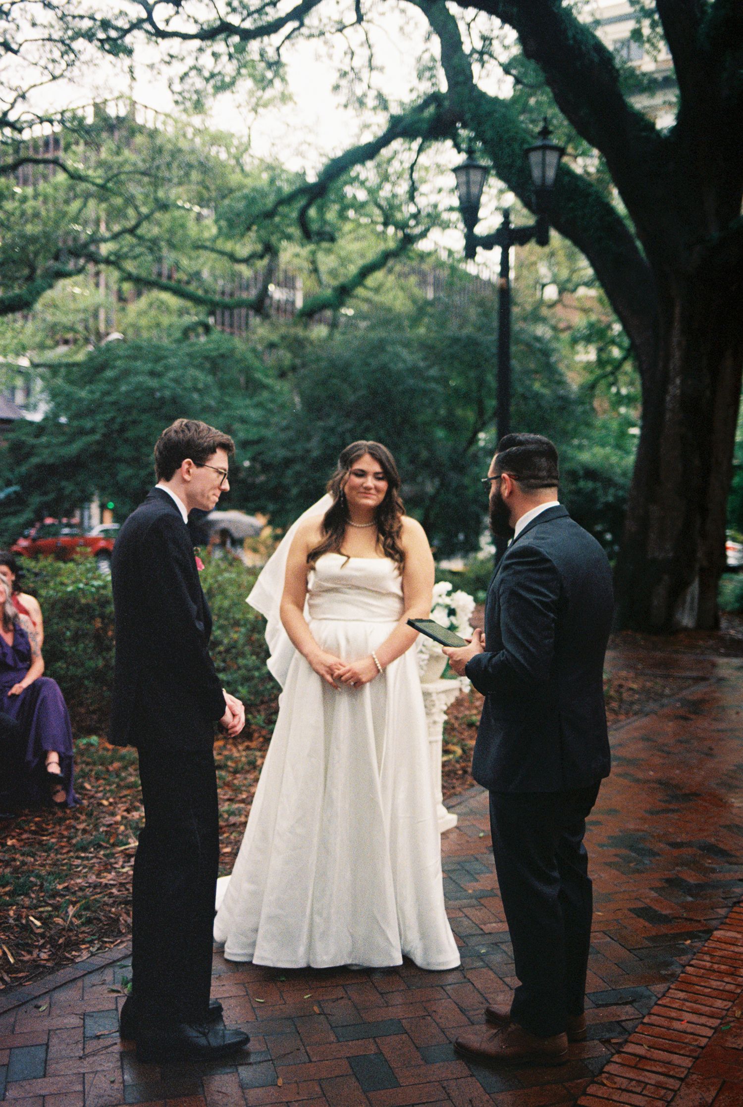 Outdoor wedding ceremony on brick path under oak trees, bride in white gown with groom and officiant.