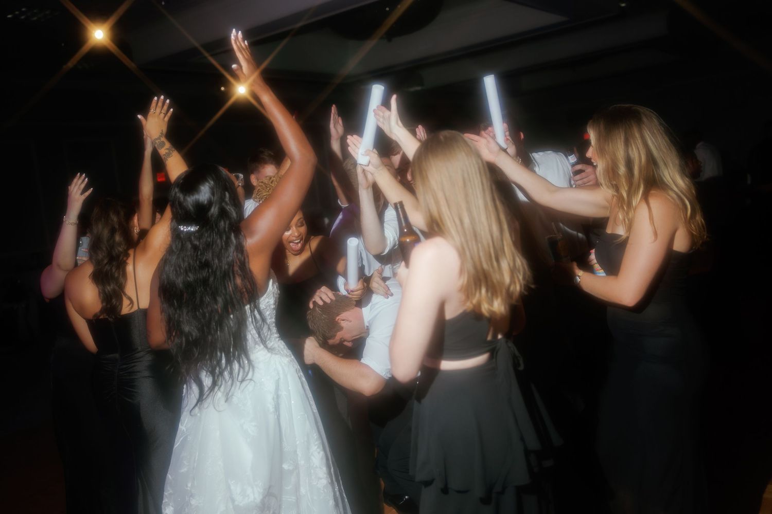 Group of women dancing at a wedding reception, holding glow sticks in a dark venue.