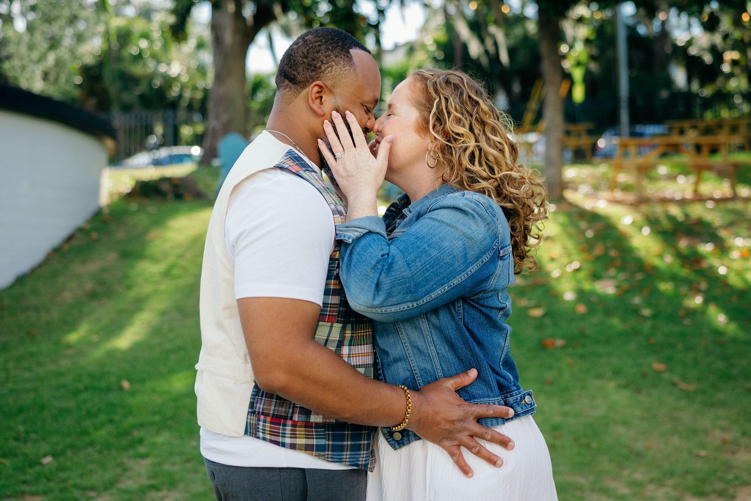 An intimate couple embraces and kisses in a sunny park setting with green trees and grass in the background.