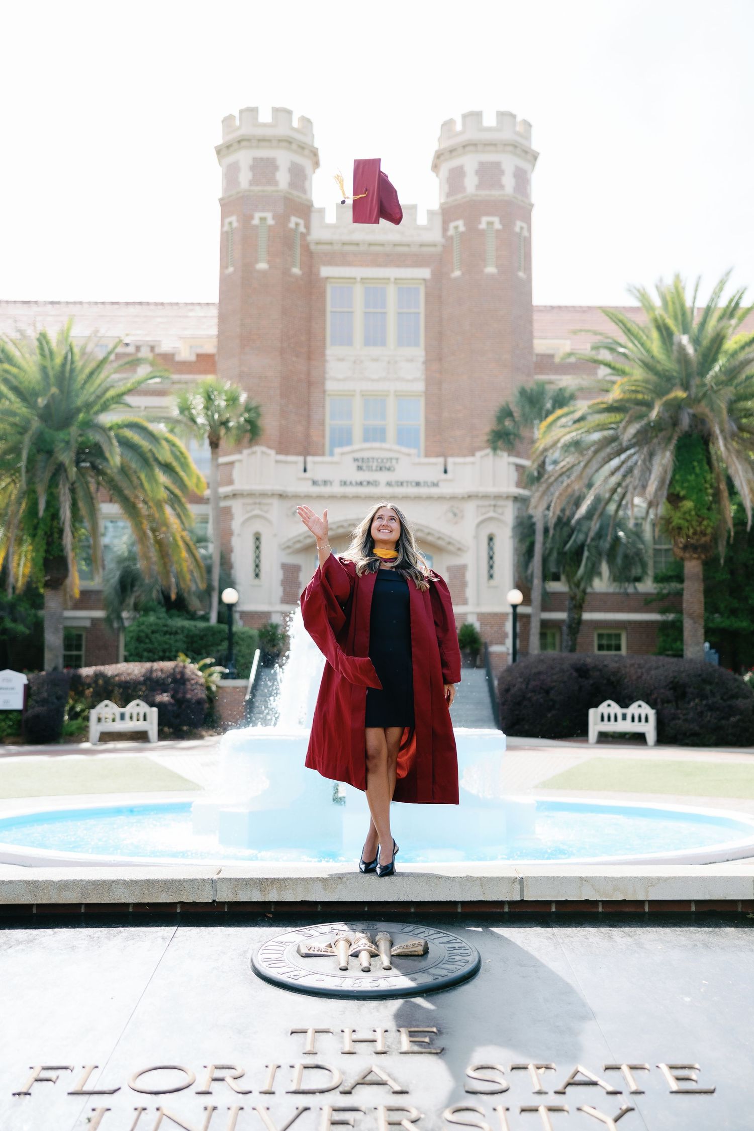 FSU graduate in maroon cap and gown poses at The Florida State University fountain outside Westcott Building.