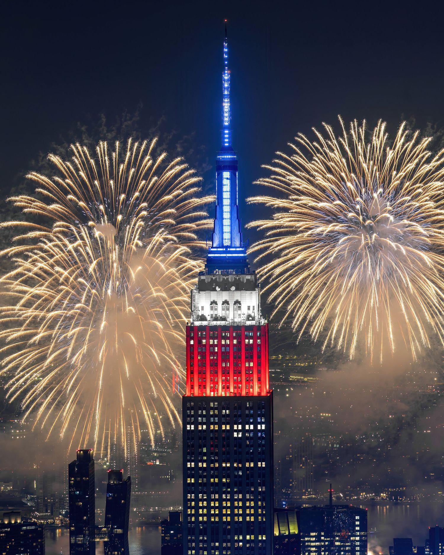 Empire State Building lit in red, white, and blue with fireworks exploding on both sides during nighttime celebration.