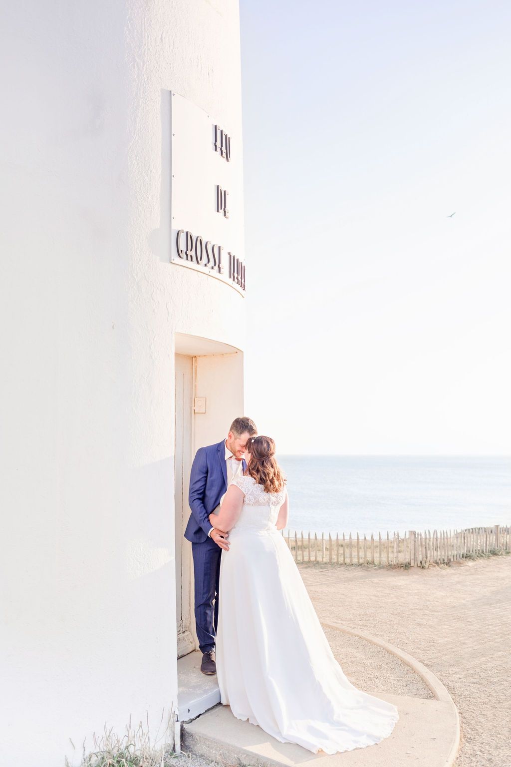 A romantic wedding couple poses against a white lighthouse wall overlooking the ocean.