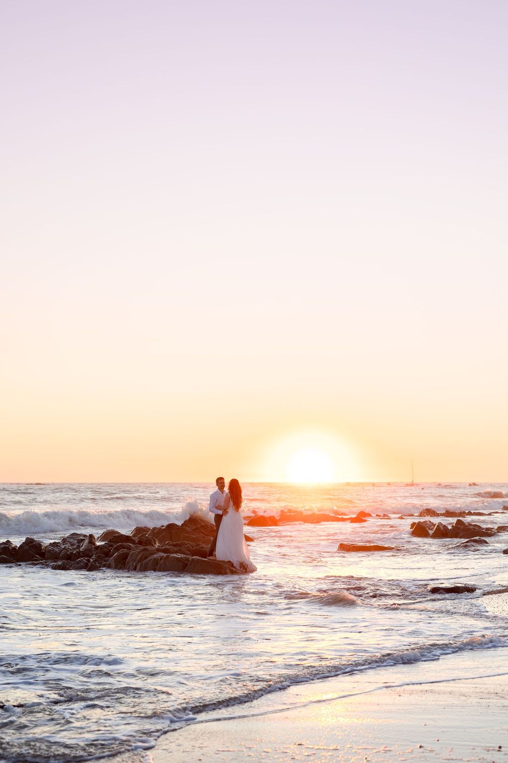 Two figures stand on rocky outcrop during vibrant ocean sunset.