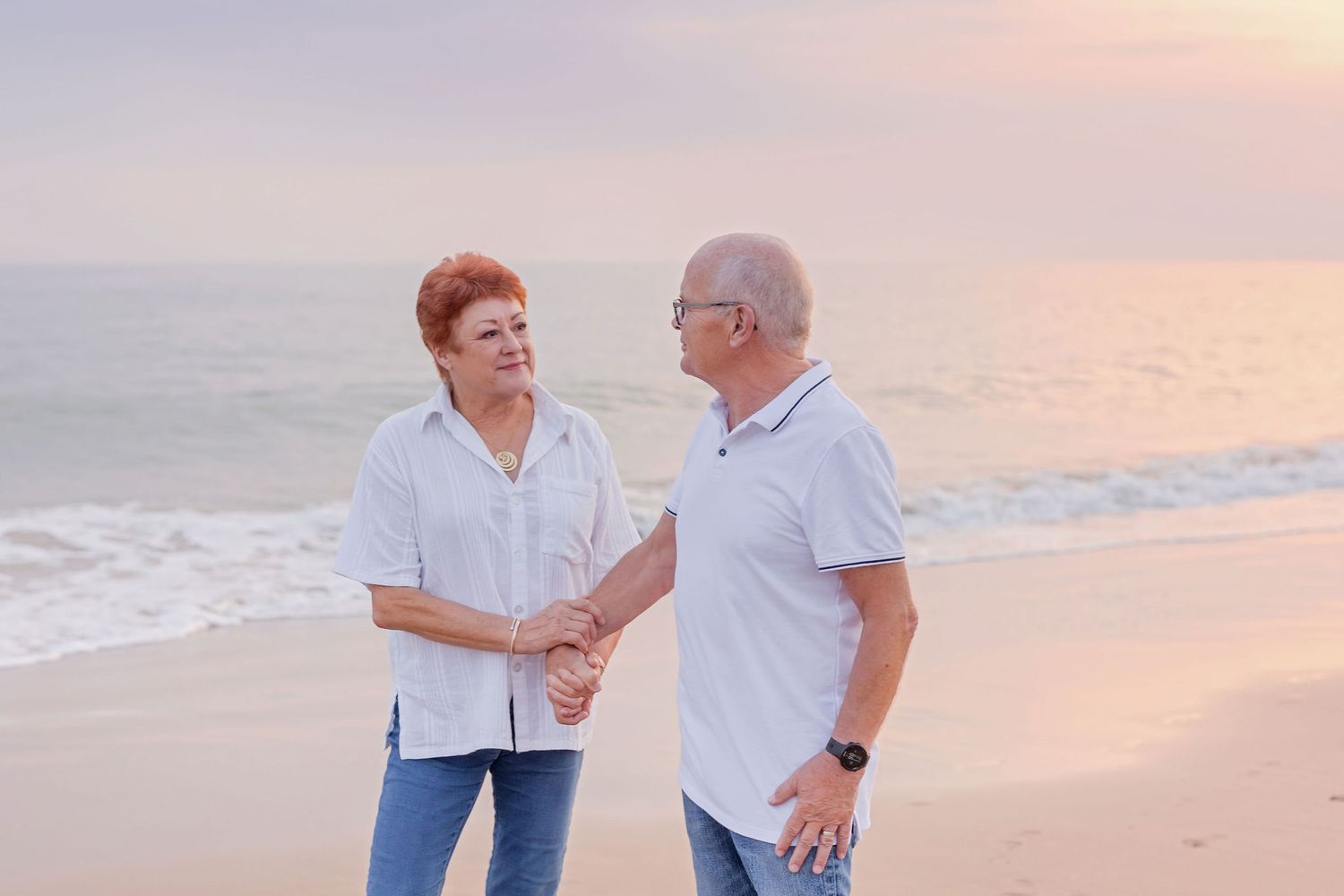 Two people sharing a handshake on beach at sunset wearing light colored shirts.