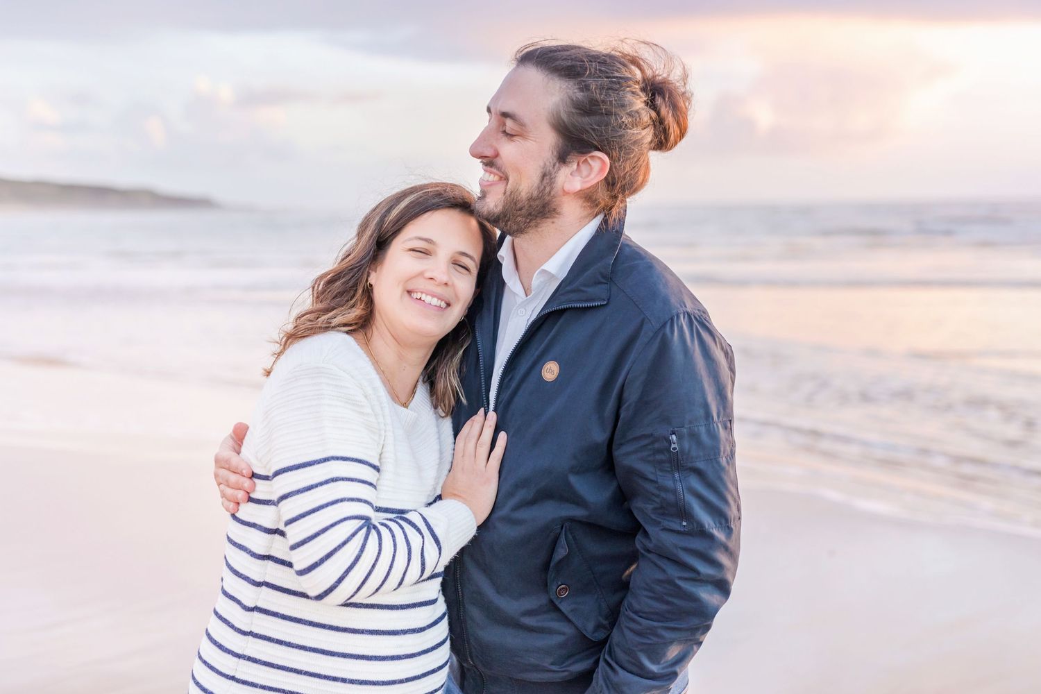 A couple embraces on the beach at sunset wearing a navy jacket and striped sweater.