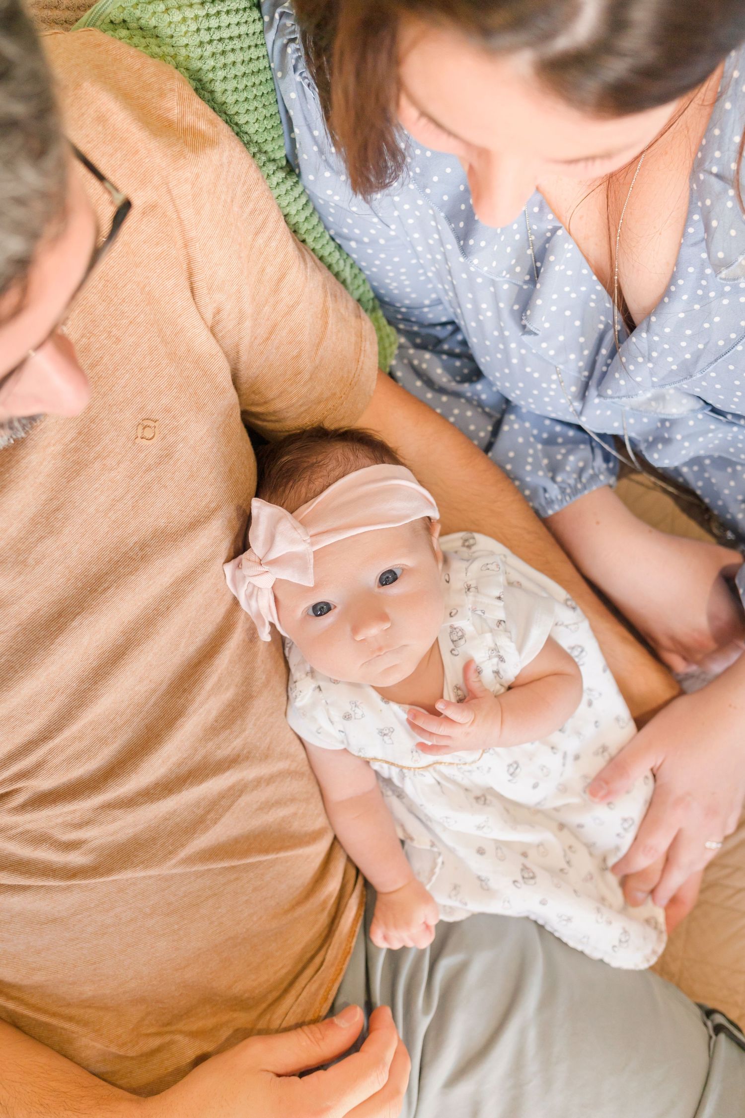 Baby in white dress lies peacefully on brown couch while being gently held.