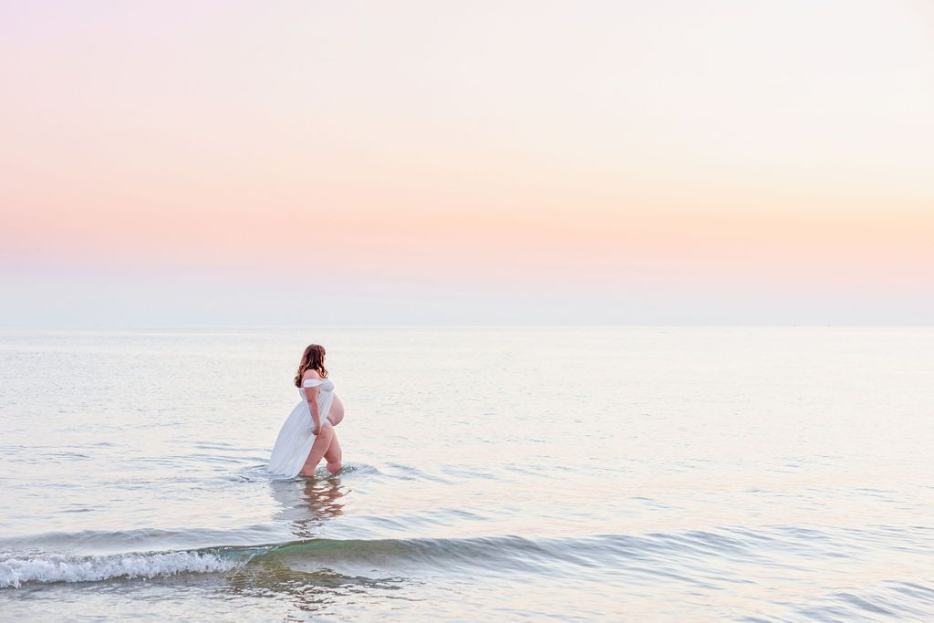 Dreamy beach portrait of woman in white dress wading through ocean at sunset.