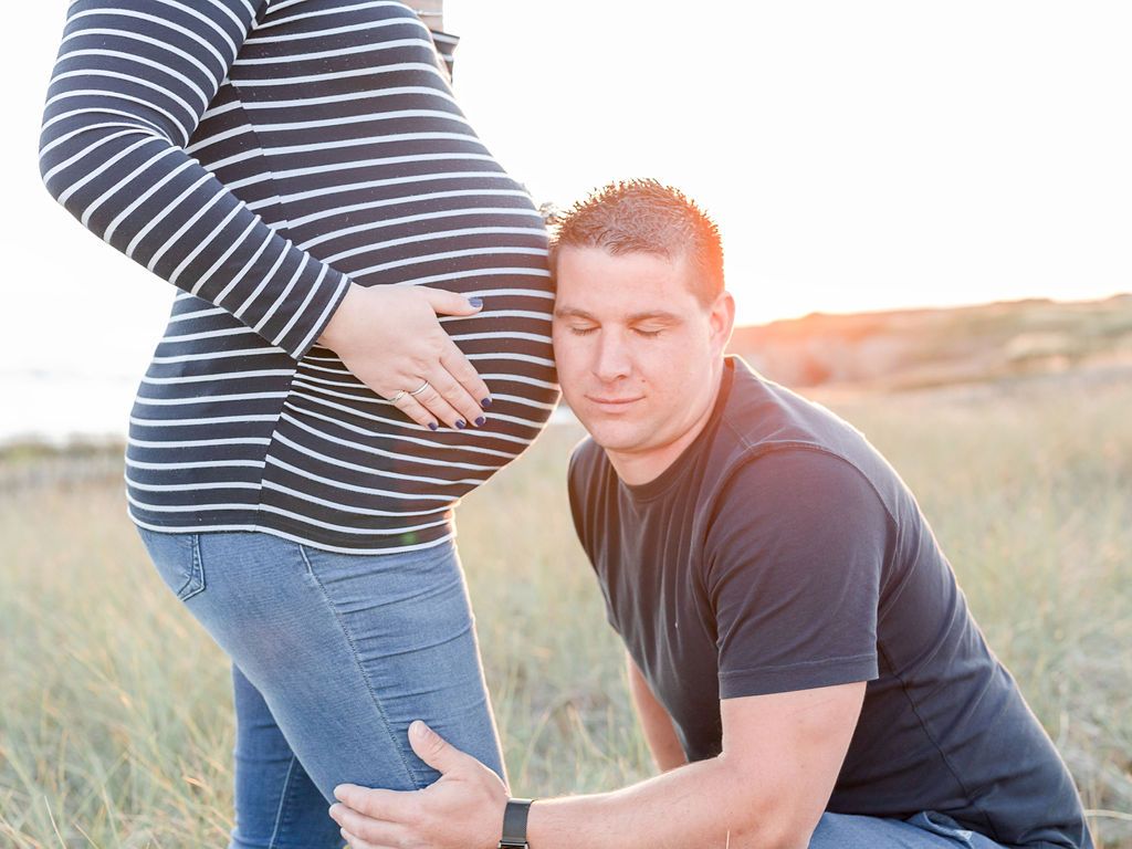 Tender maternity moment captured in seaside grass during golden hour.