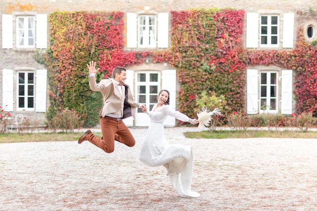 A couple in wedding attire leaps joyfully in front of an ivy-covered estate building.