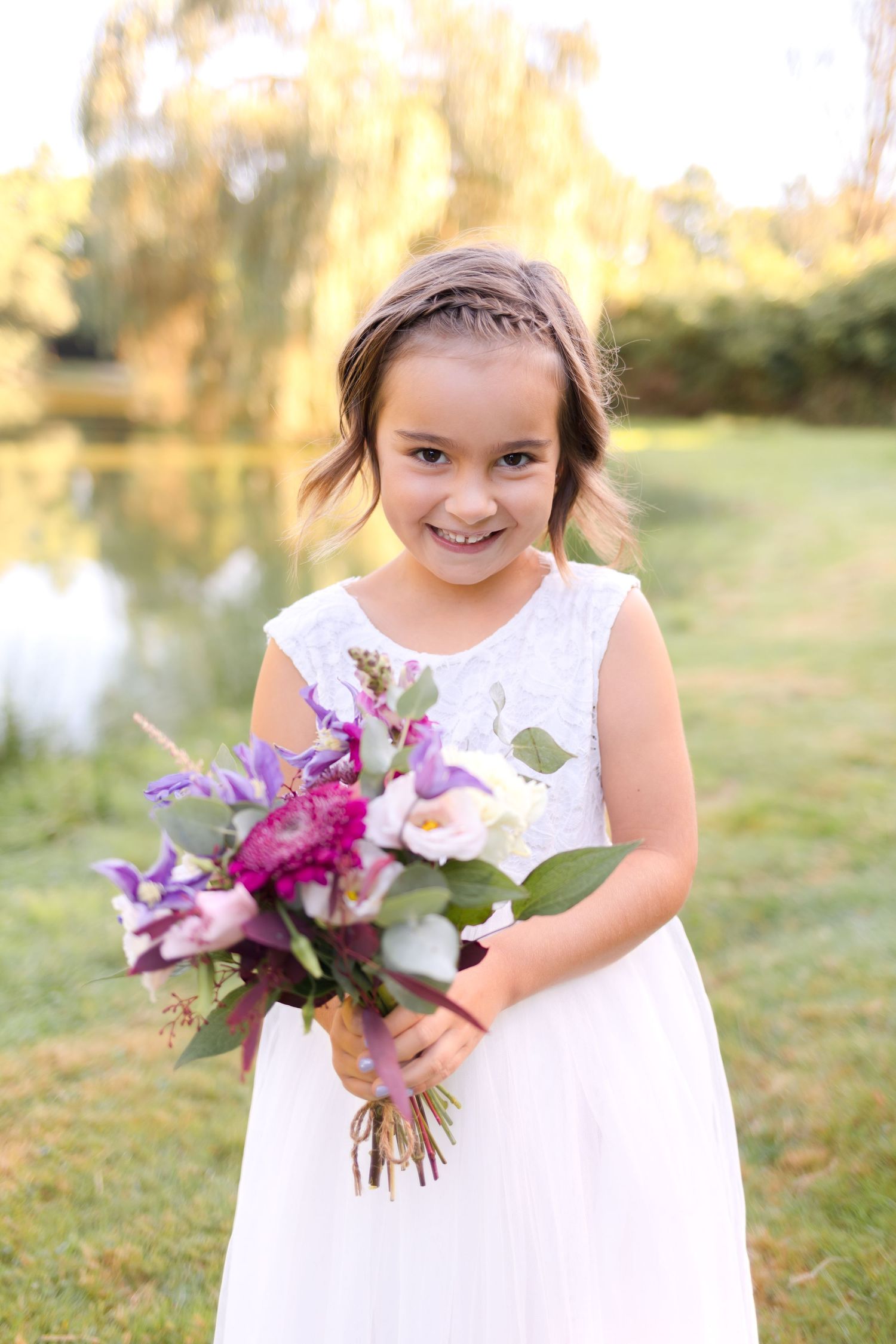 Young attendant in a white dress holding a vibrant purple and pink bouquet by a pond.