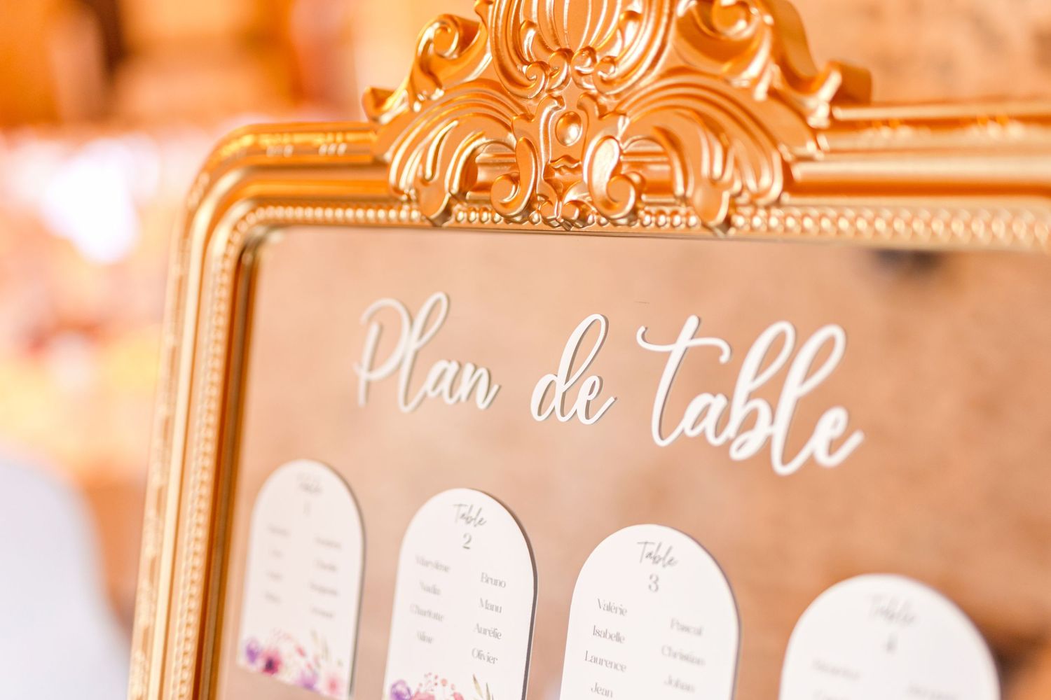 'Plan de Table' written in white script on an ornate gold frame displaying seating assignments.