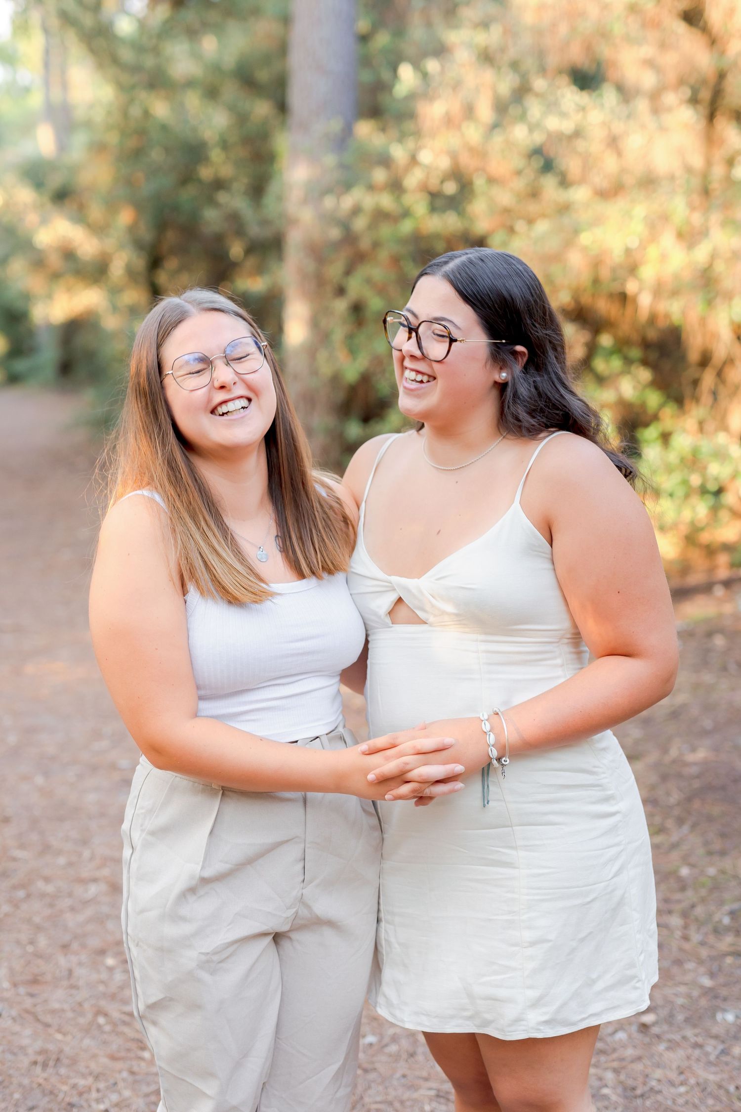 Two friends in matching white outfits sharing a laugh together in a sunny forest setting.