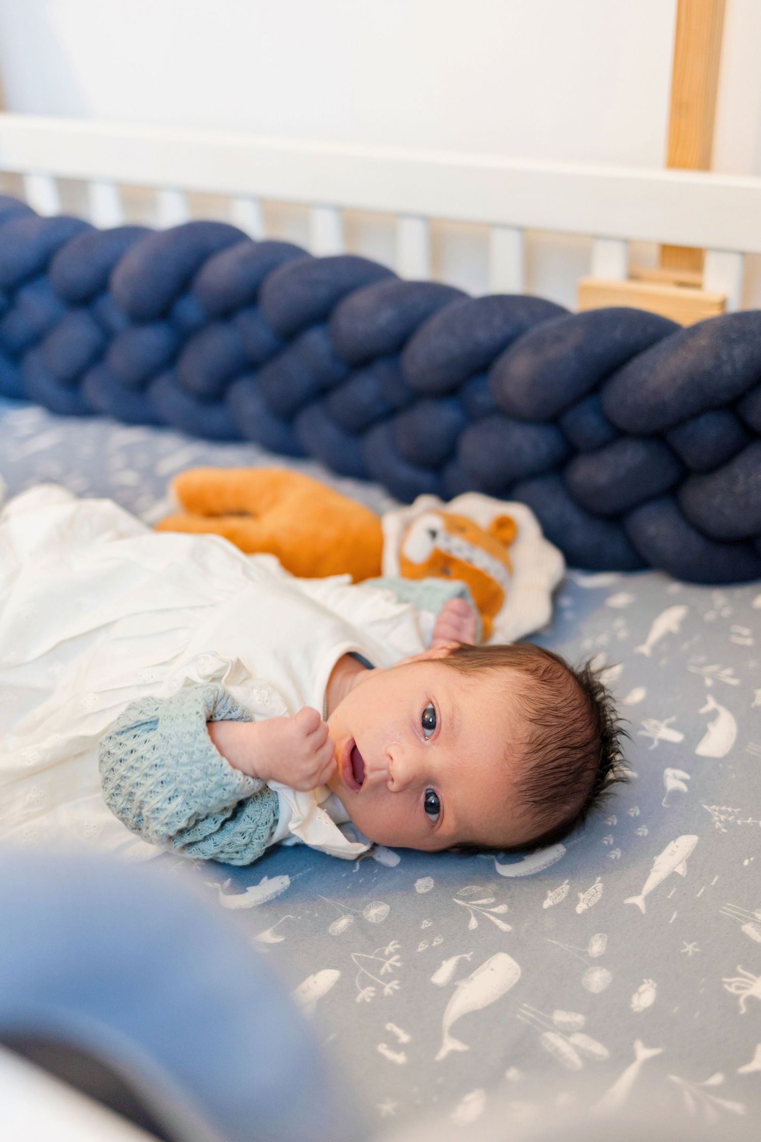 Baby in white dress and blue sweater lying on patterned bedding with navy braided crib bumper.