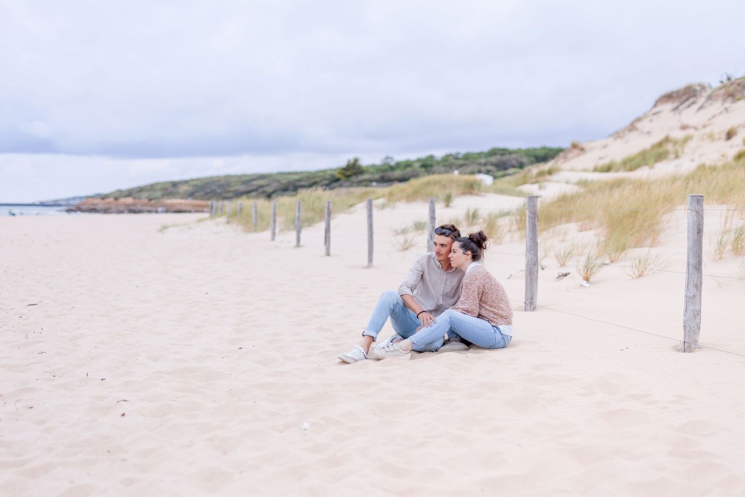 Couple sitting together on sandy beach near wooden posts with coastal dunes in background.
