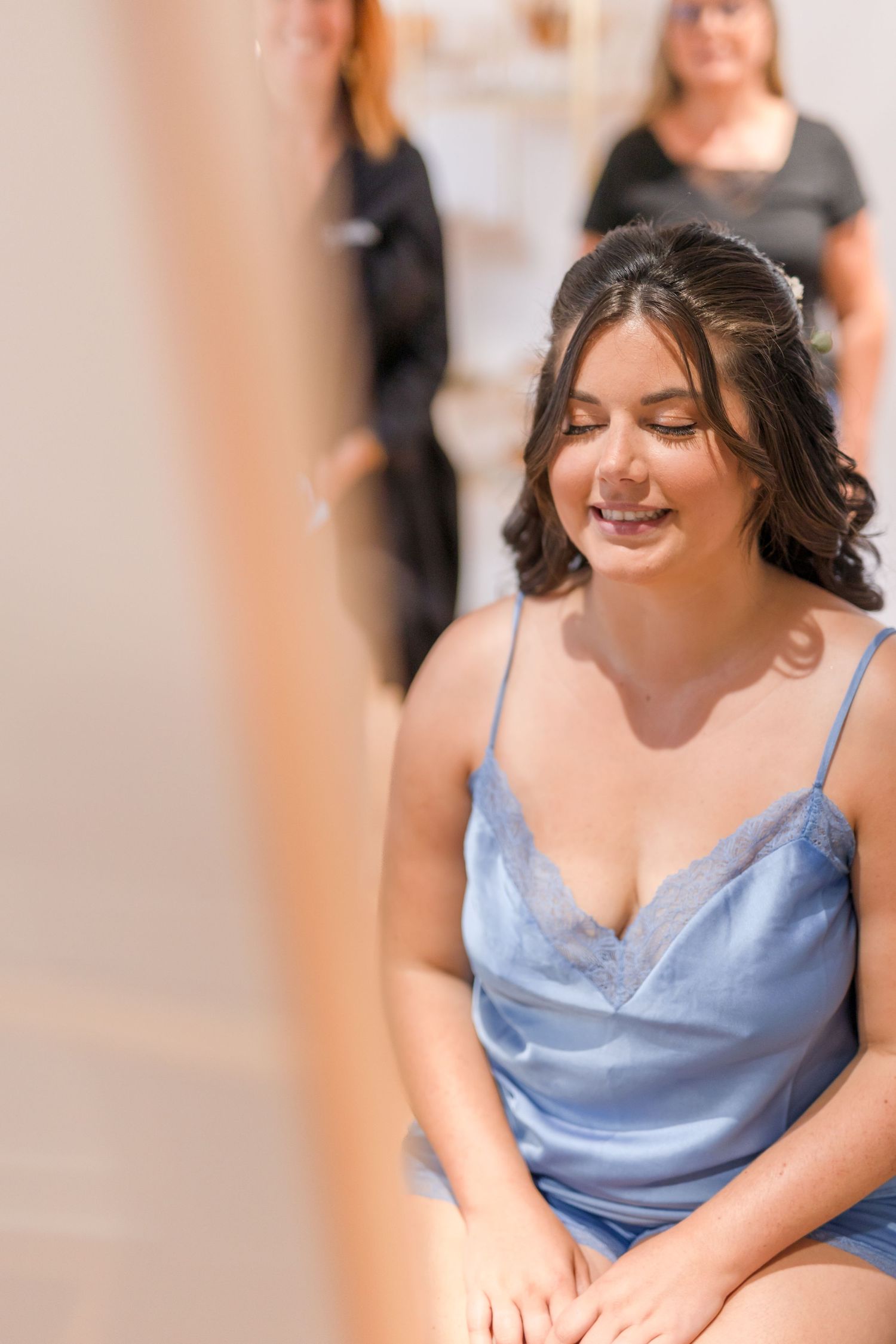 Someone in a blue satin dress smiles while seated during preparations.