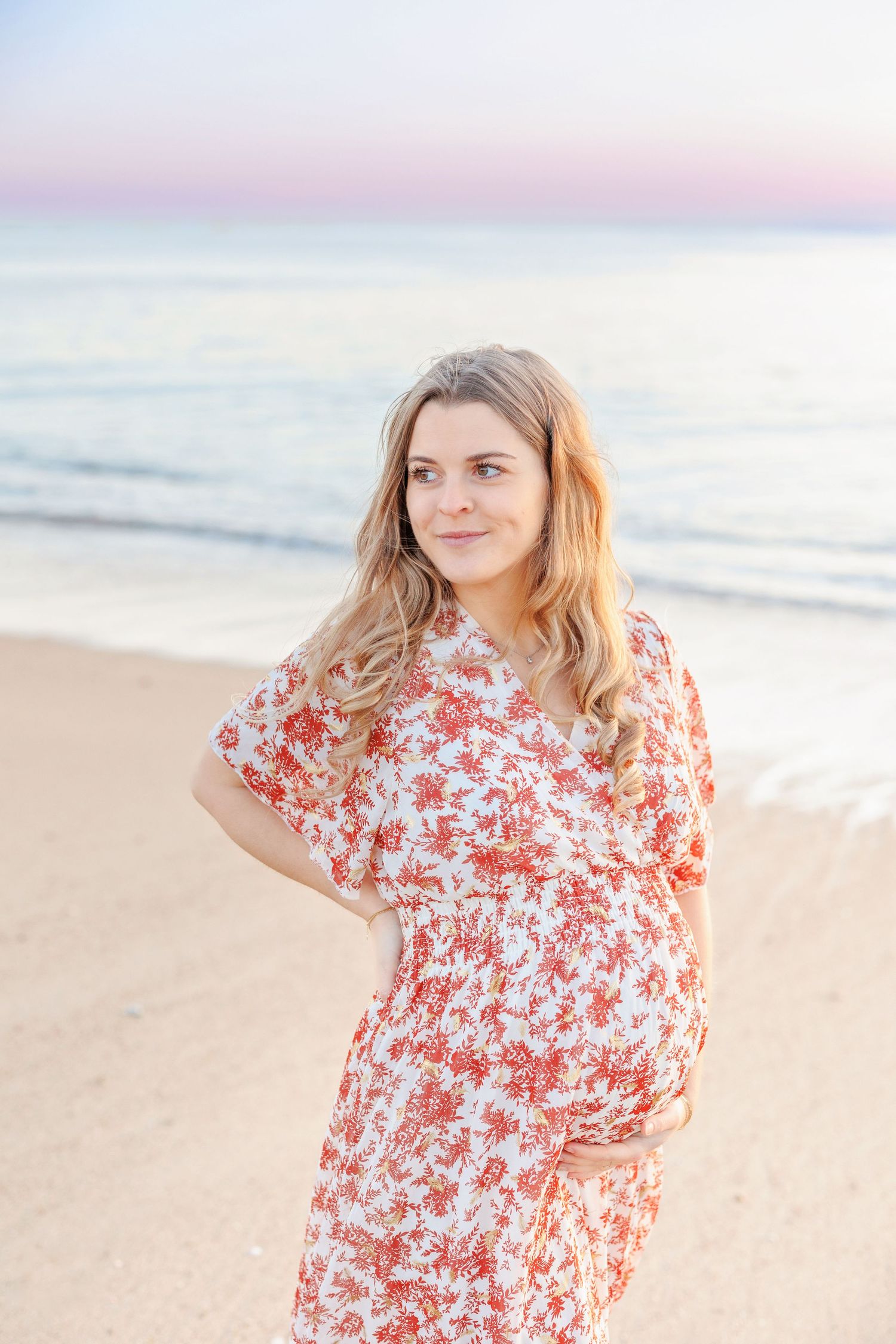 A person in a red and white floral dress poses on the beach during sunset.