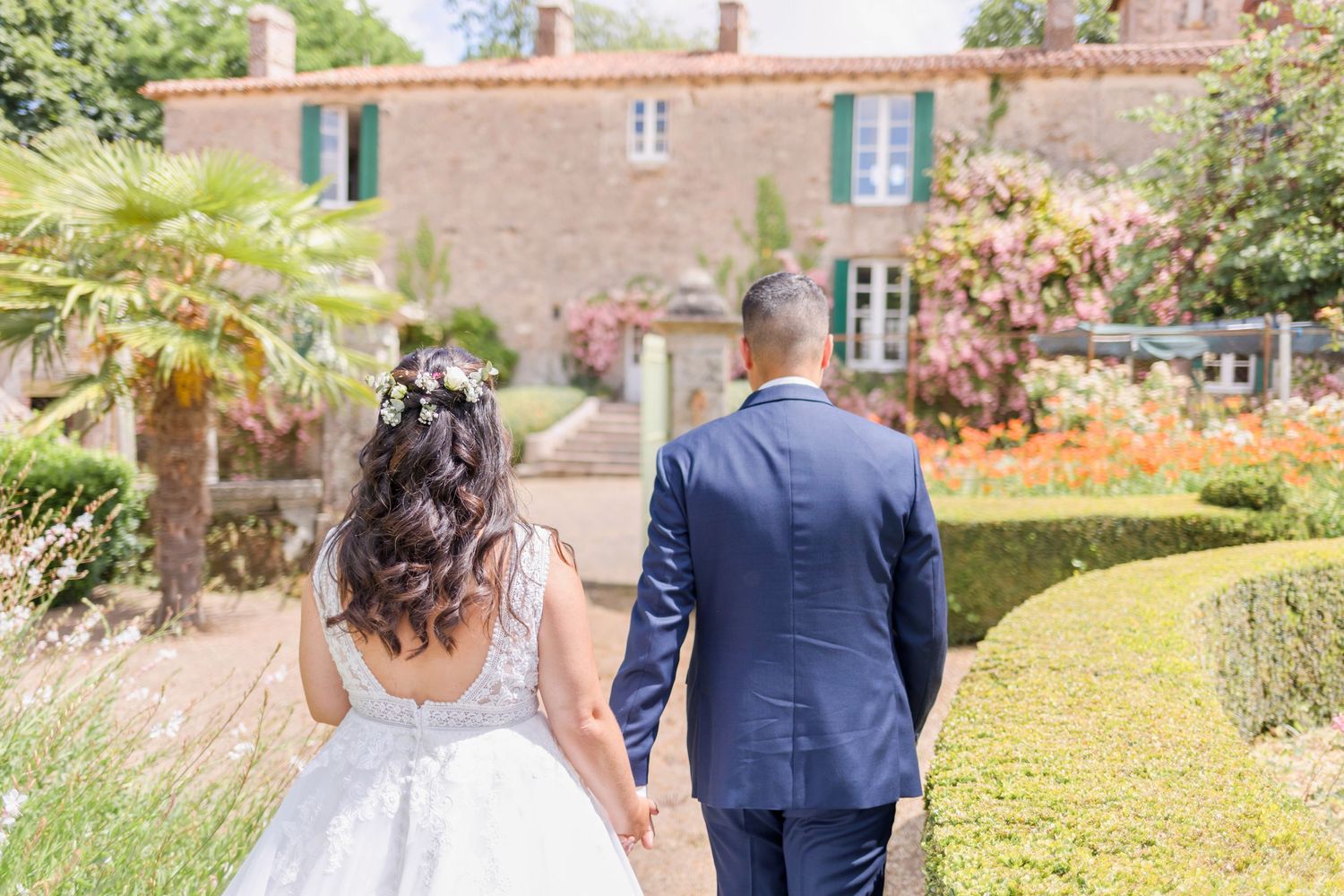 A wedding couple walks hand in hand through a manicured garden with a stone building in the background.