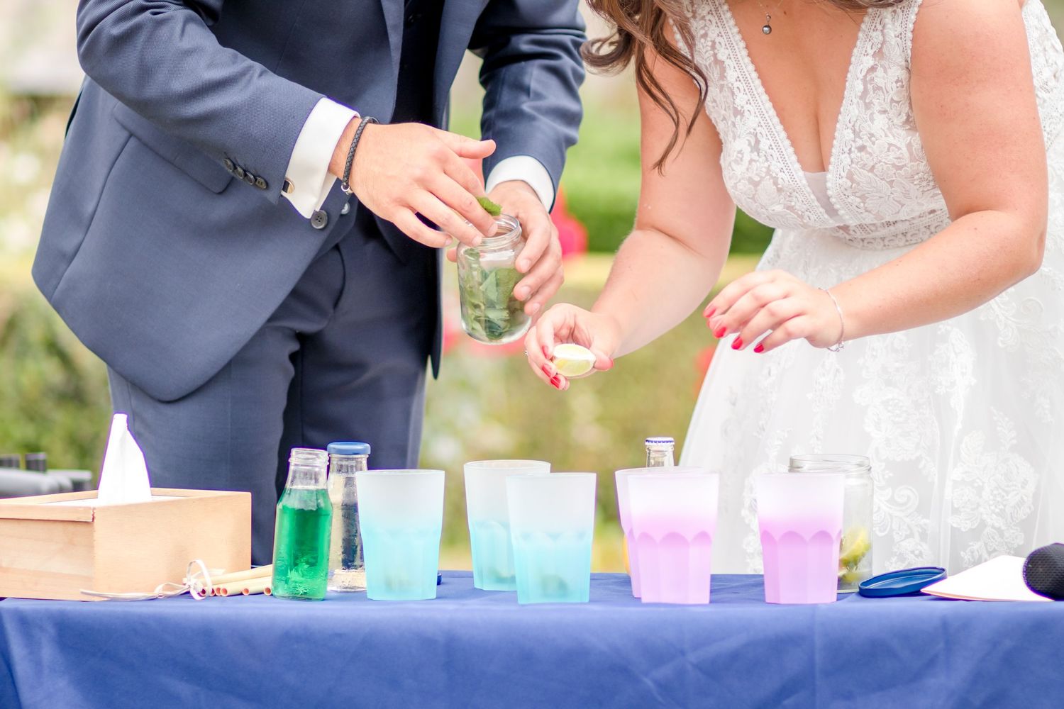 Close-up of hands pouring bright colored liquids into small glasses on a navy blue tablecloth.