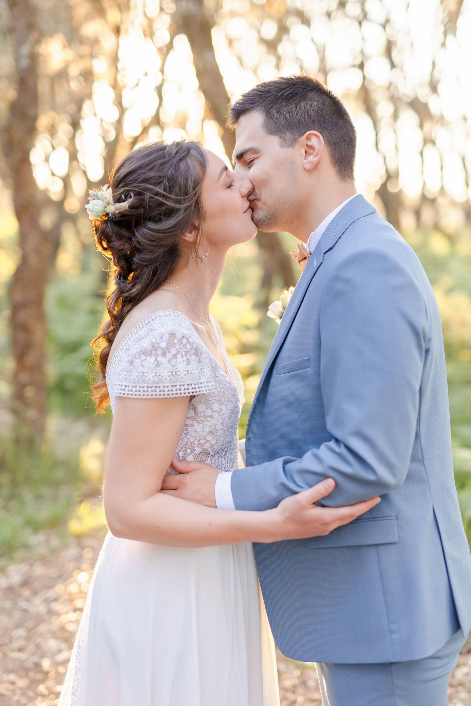 A romantic wedding kiss captured in a sun-dappled forest setting.