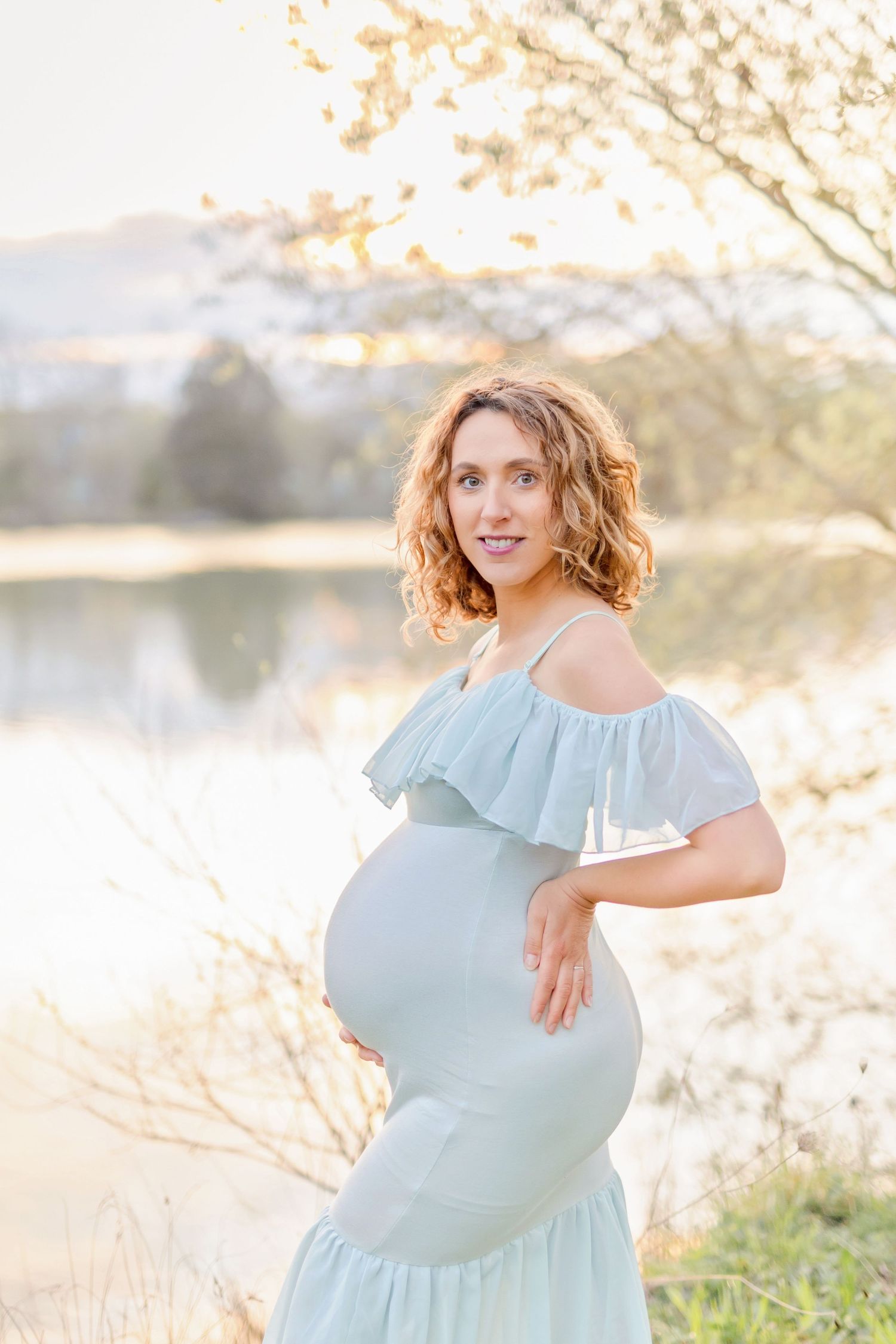 Serene maternity portrait by a lake wearing an elegant blue off-shoulder gown with ruffles.