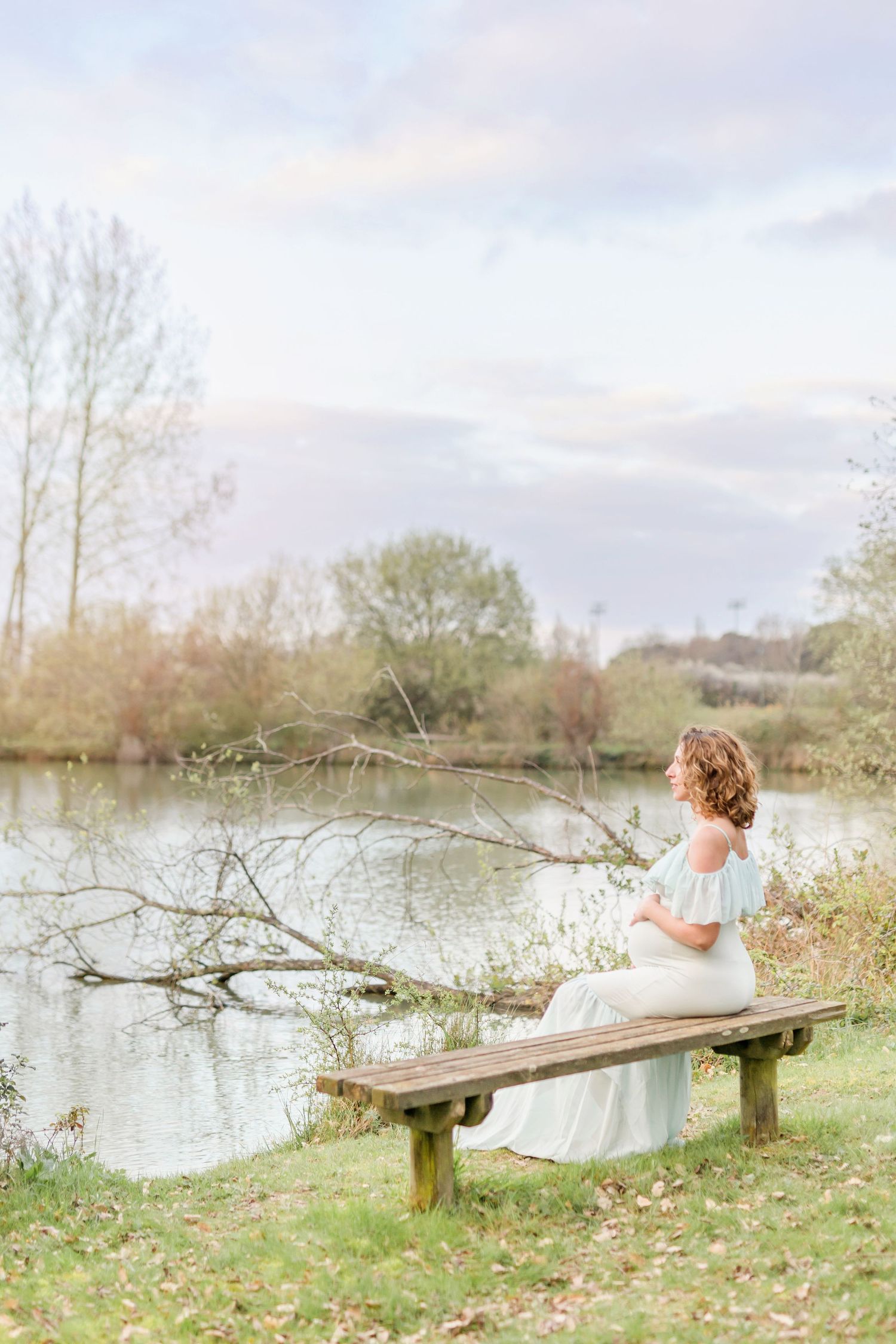 Serene lakeside maternity session on a wooden bench with wispy clouds overhead.