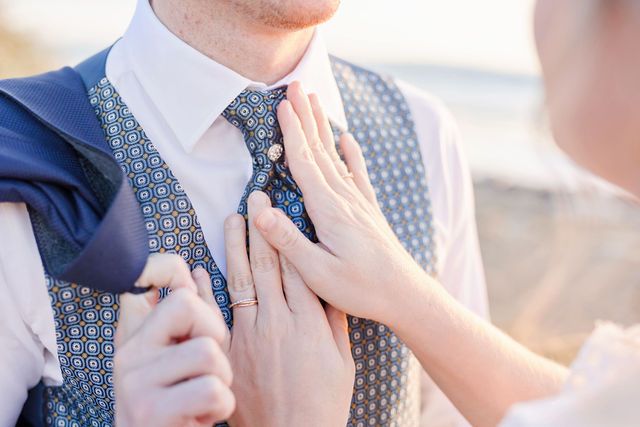 Hands adjusting groom's tie during wedding preparations on beach.