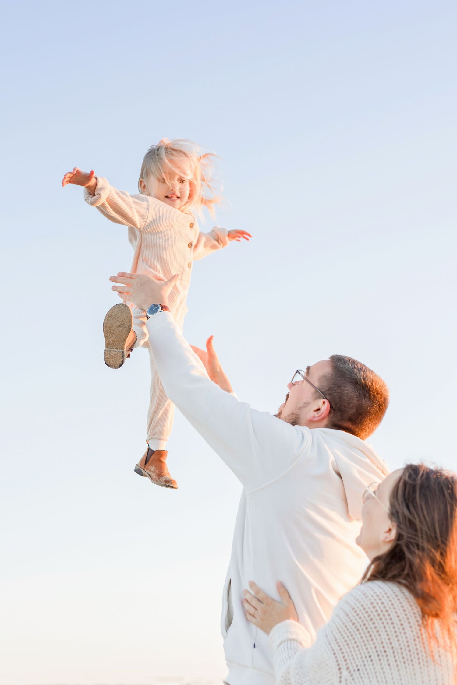 A toddler in white clothing is lifted joyfully into the air by their parents at sunset.