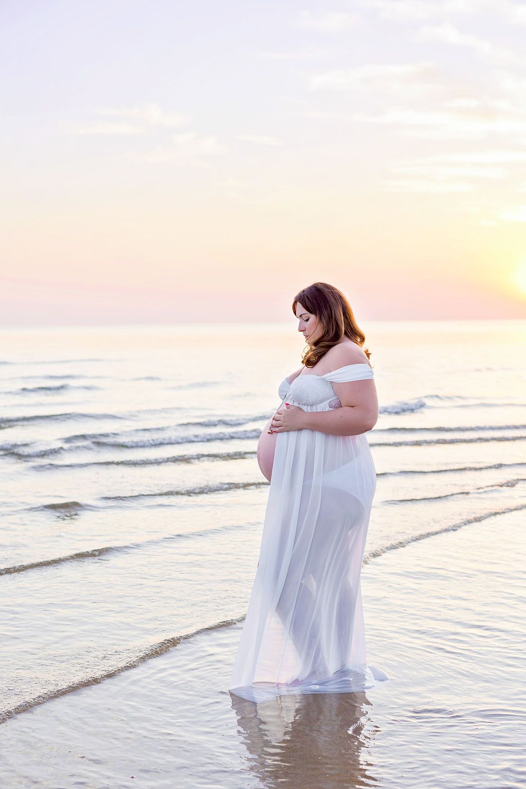 Pregnant woman in white dress poses thoughtfully by the shoreline at dusk.
