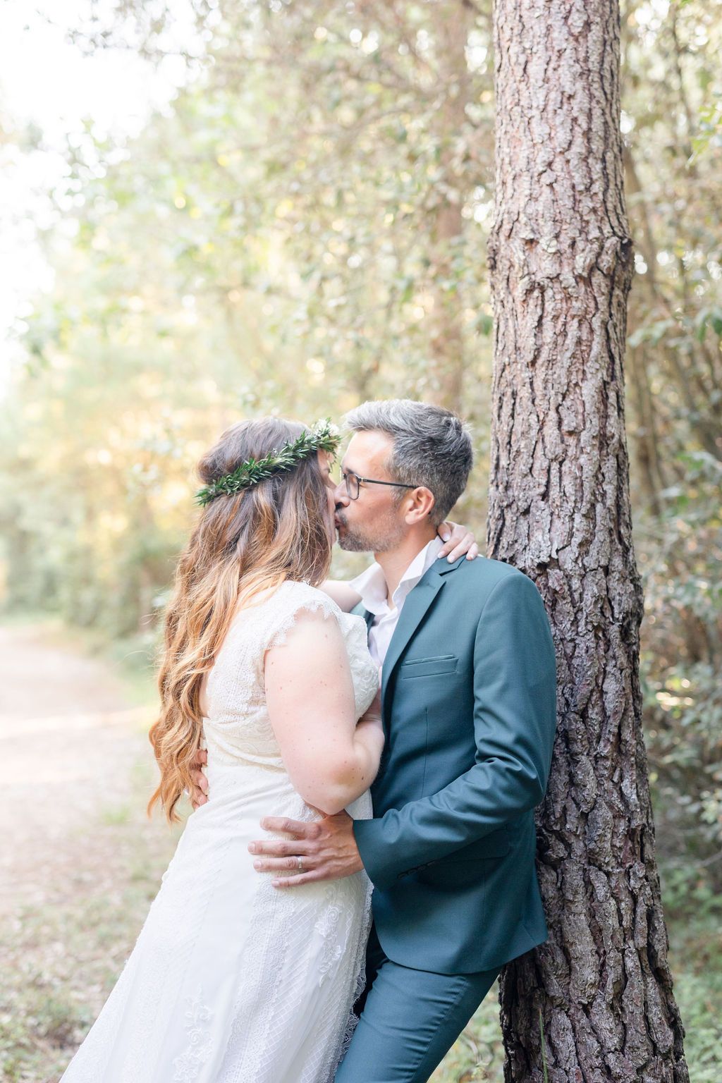 Romantic wedding couple embraces against a tree trunk in a sunlit forest path.
