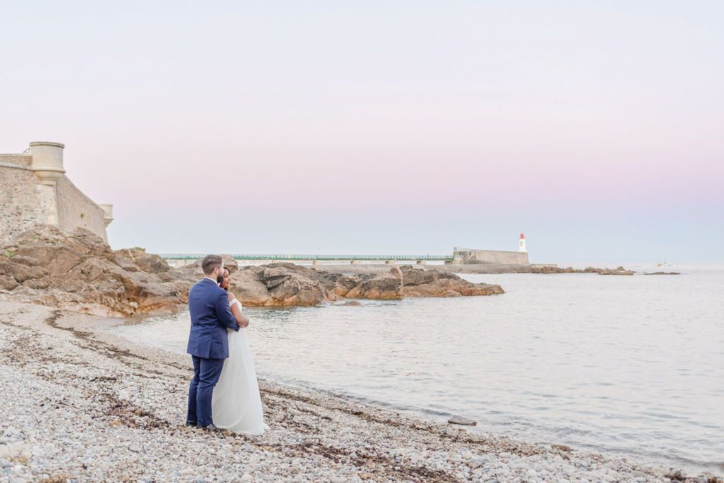 Wedding couple stands on rocky shoreline with lighthouse and ocean in background at dusk.