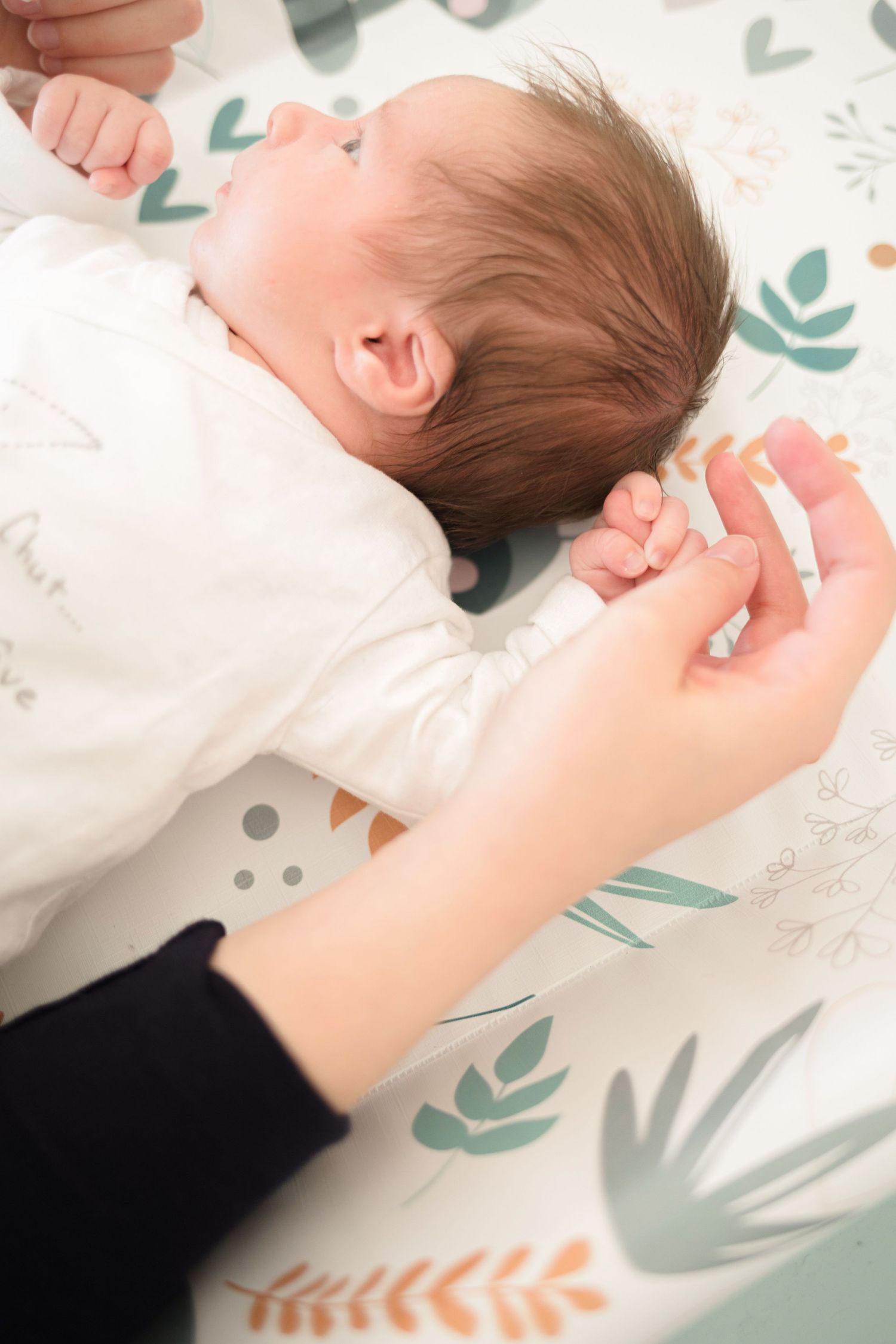 Gentle hands reach to comfort baby lying on nature-patterned blanket.