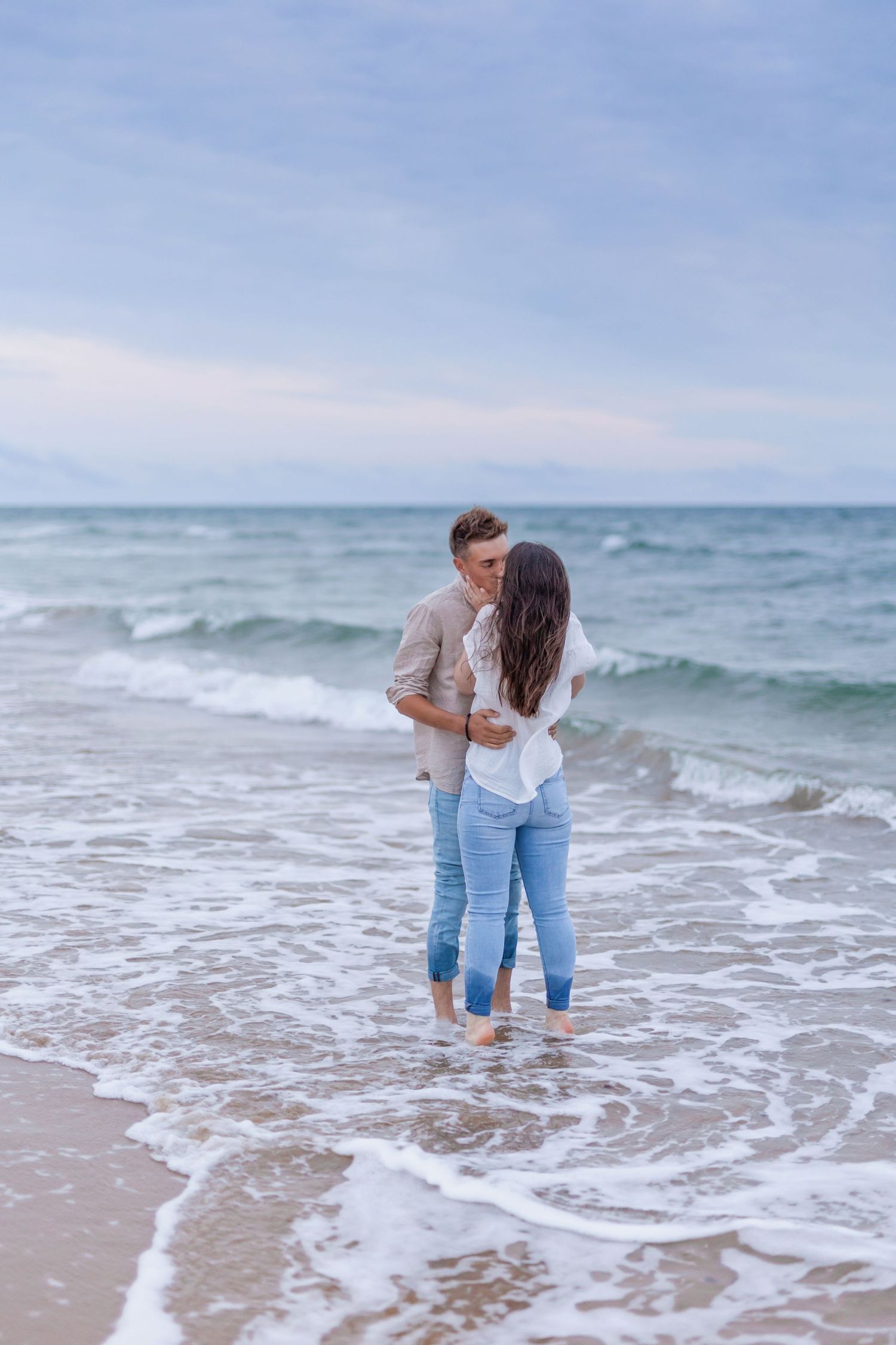 A couple standing in the shallow ocean waves on a cloudy beach day.