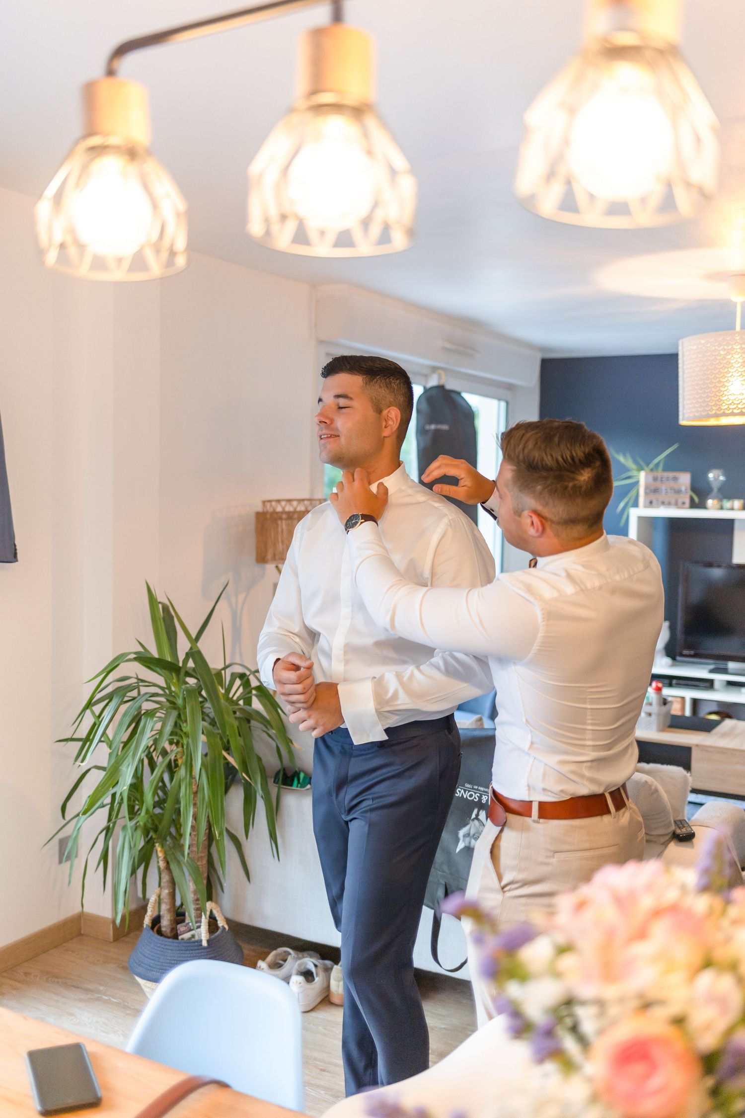 Wedding preparations showing men getting dressed under pendant lights with indoor plants in background.