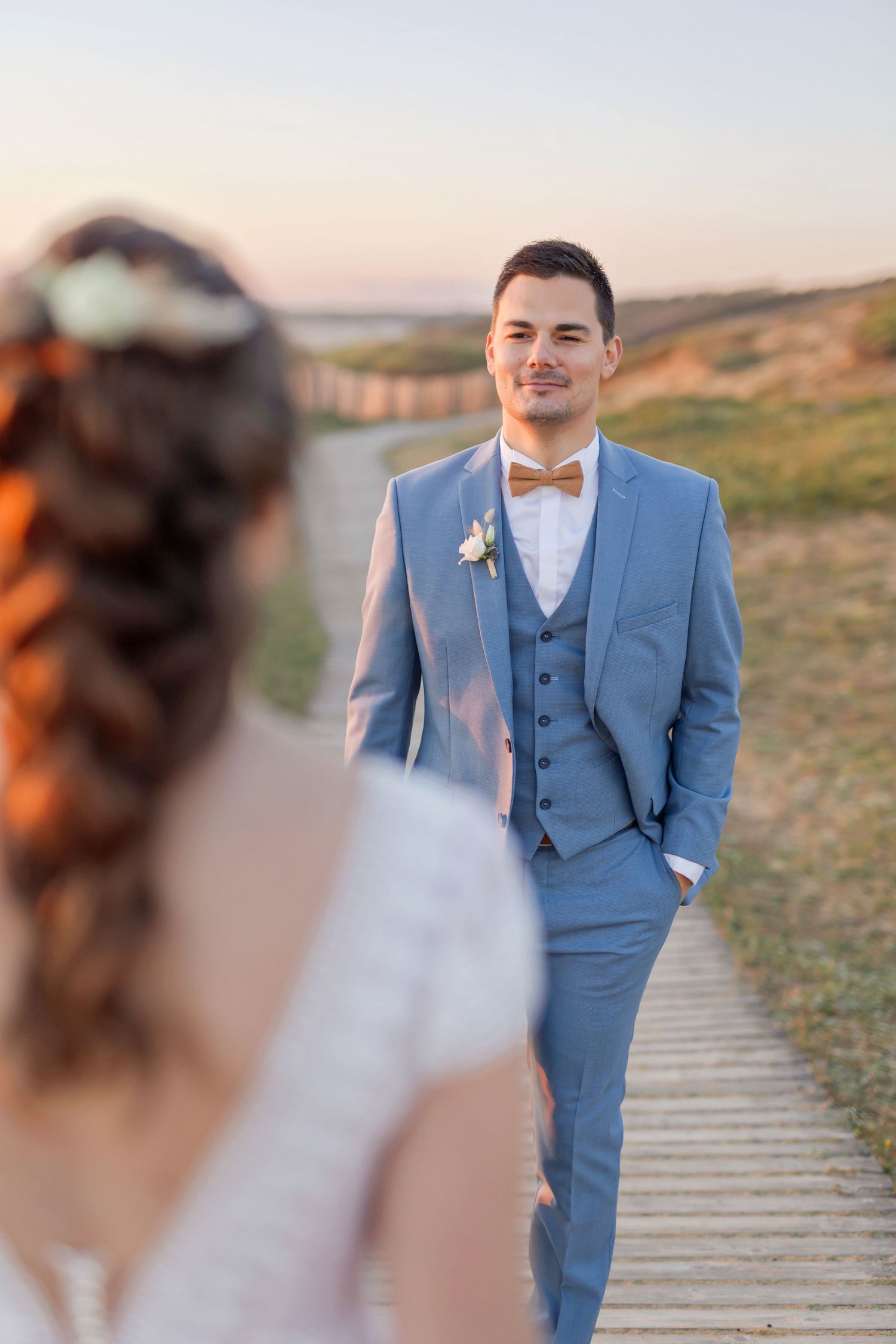 Groom in light blue suit stands on wooden boardwalk during sunset photoshoot.