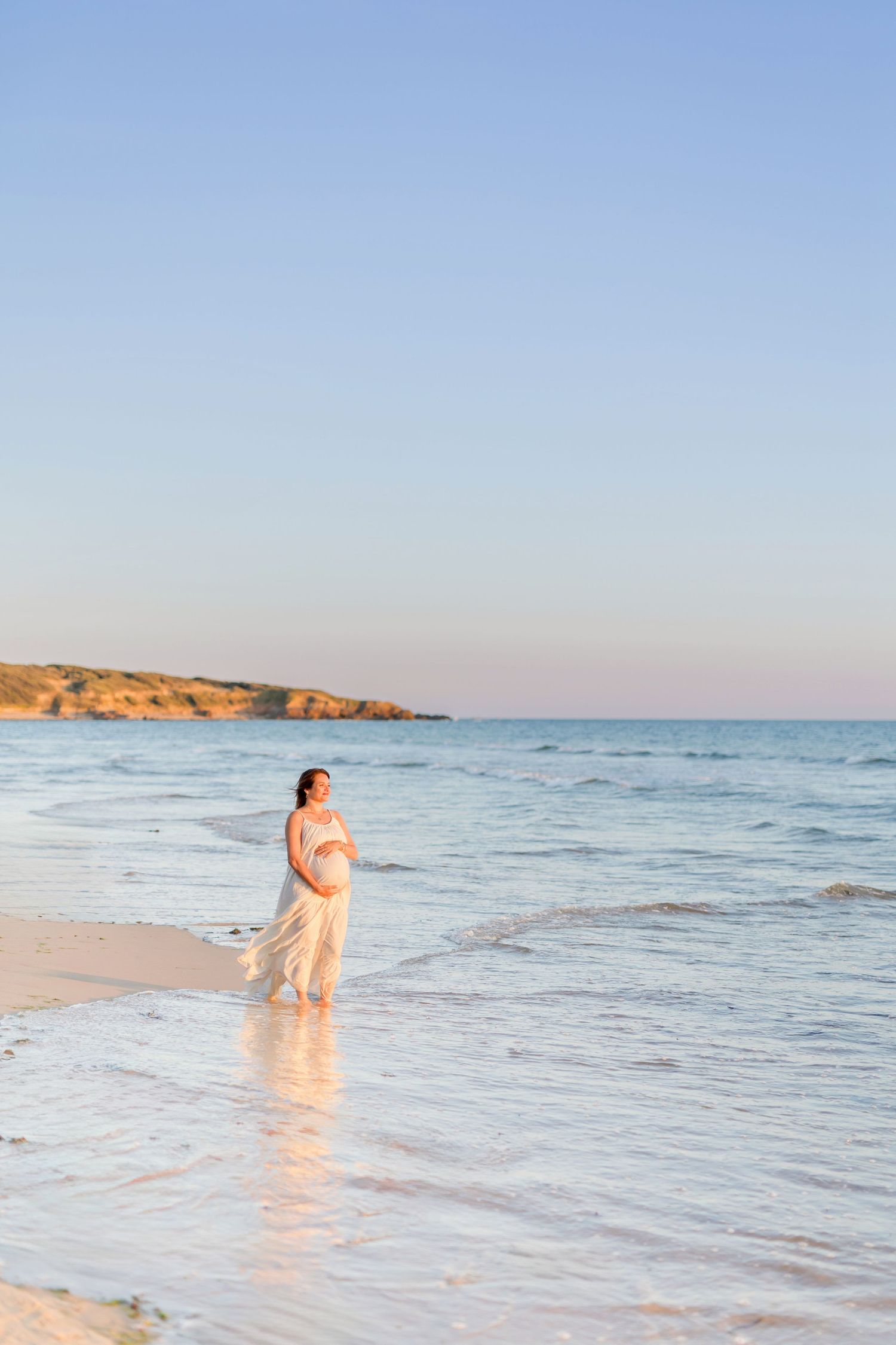 Person in flowing dress stands in gentle waves on sandy beach at dusk.