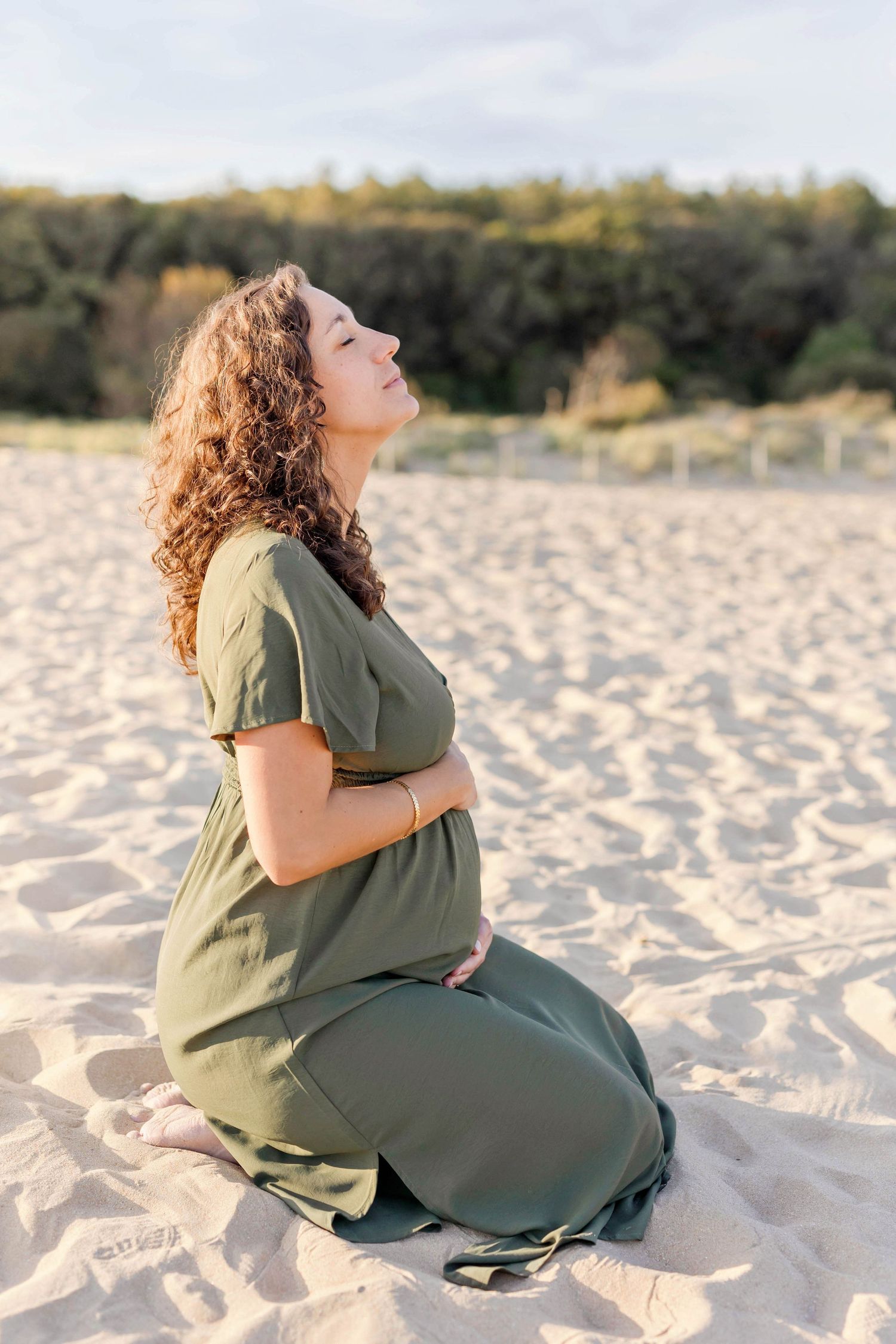 A pregnant woman in green dress sits contemplatively on sandy beach during golden hour.