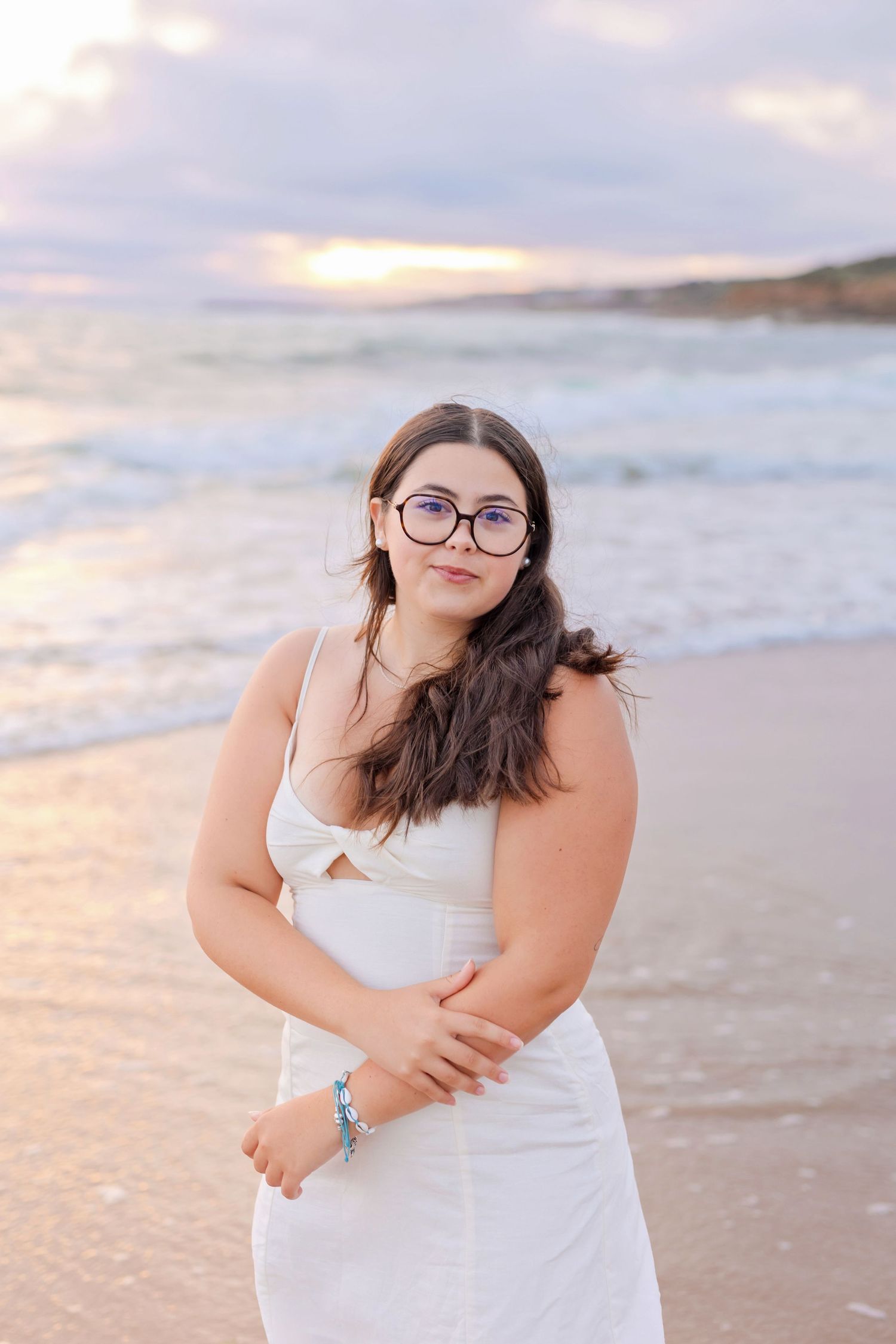 A person in a white dress and glasses poses on the beach during golden hour.