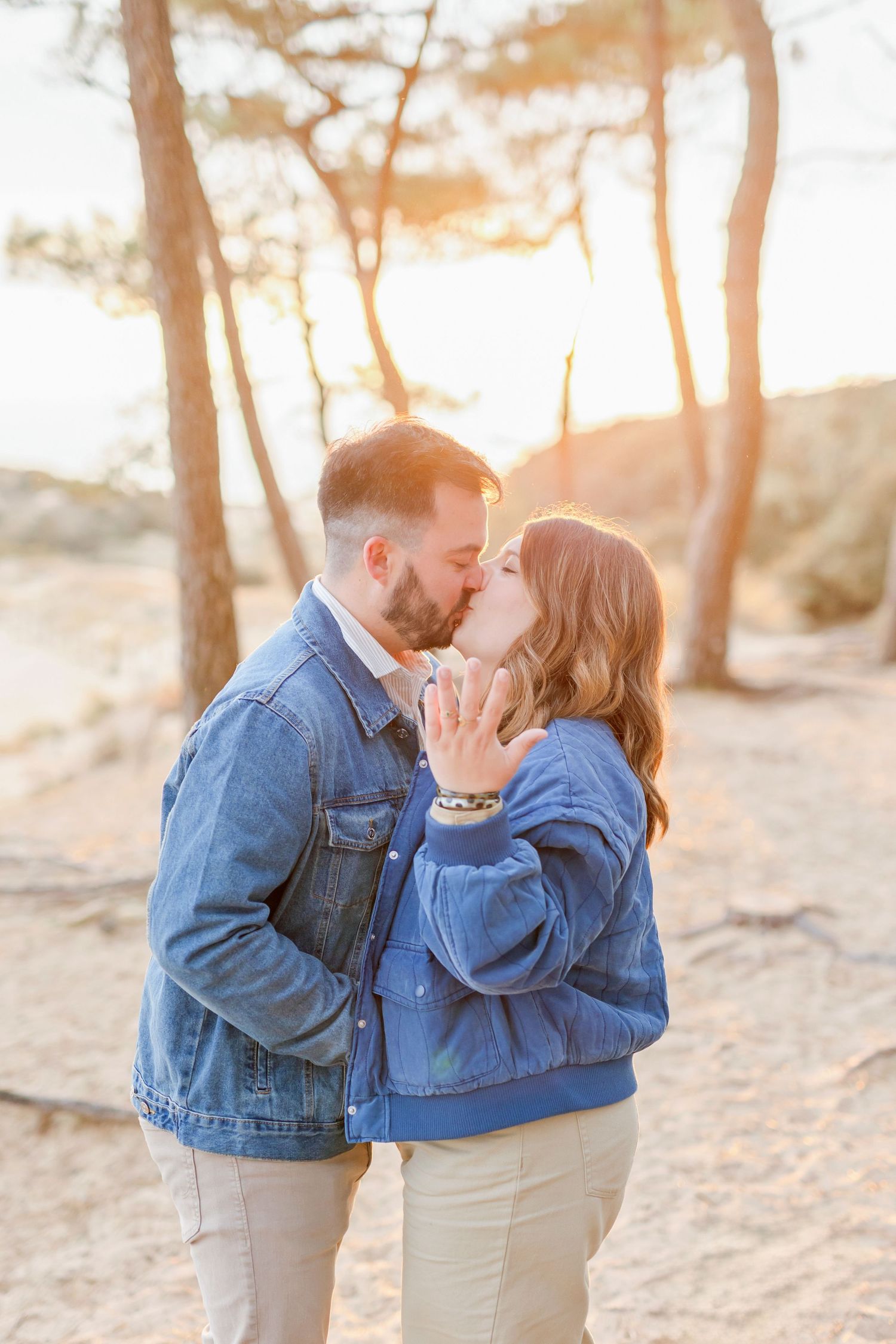 A gentle kiss shared between two people in denim jackets with sunset-lit trees in background.