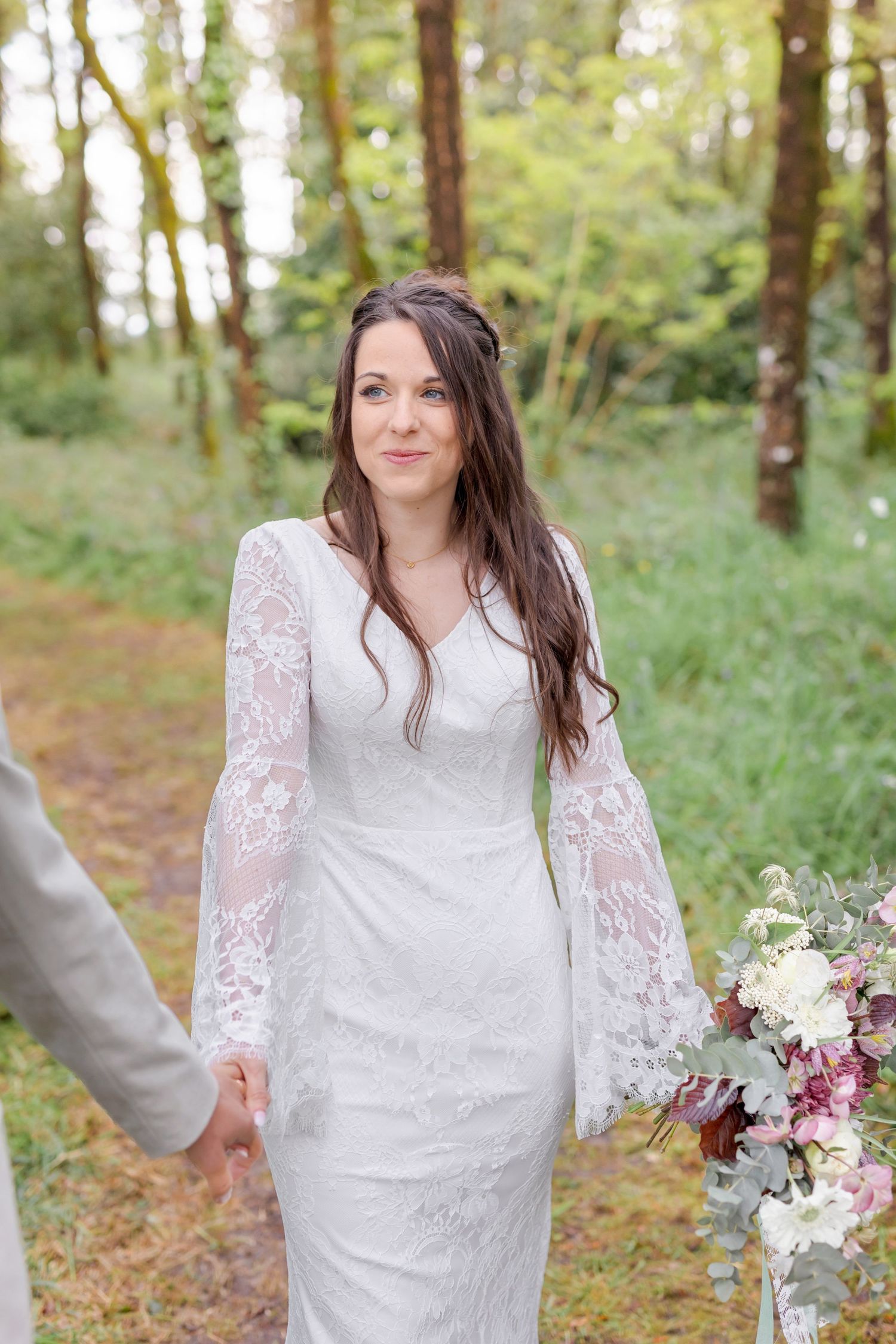 Bride wearing a white lace wedding dress stands in a sunlit forest holding a pink and white bouquet.