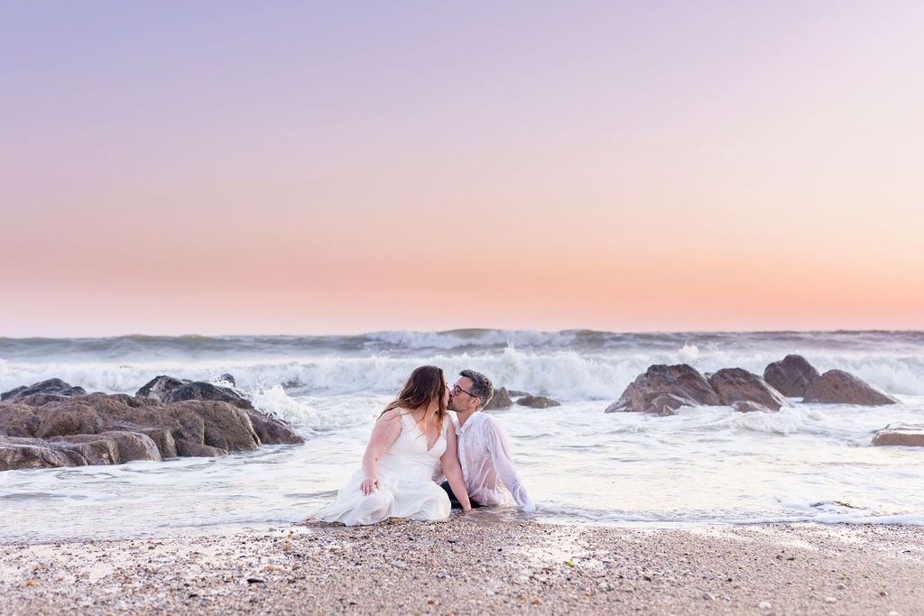 A couple sits together on beach rocks during sunset with waves crashing behind them.
