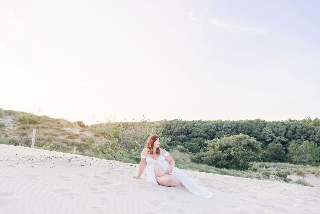 Candid maternity photo on sandy dunes with forest backdrop at golden hour.