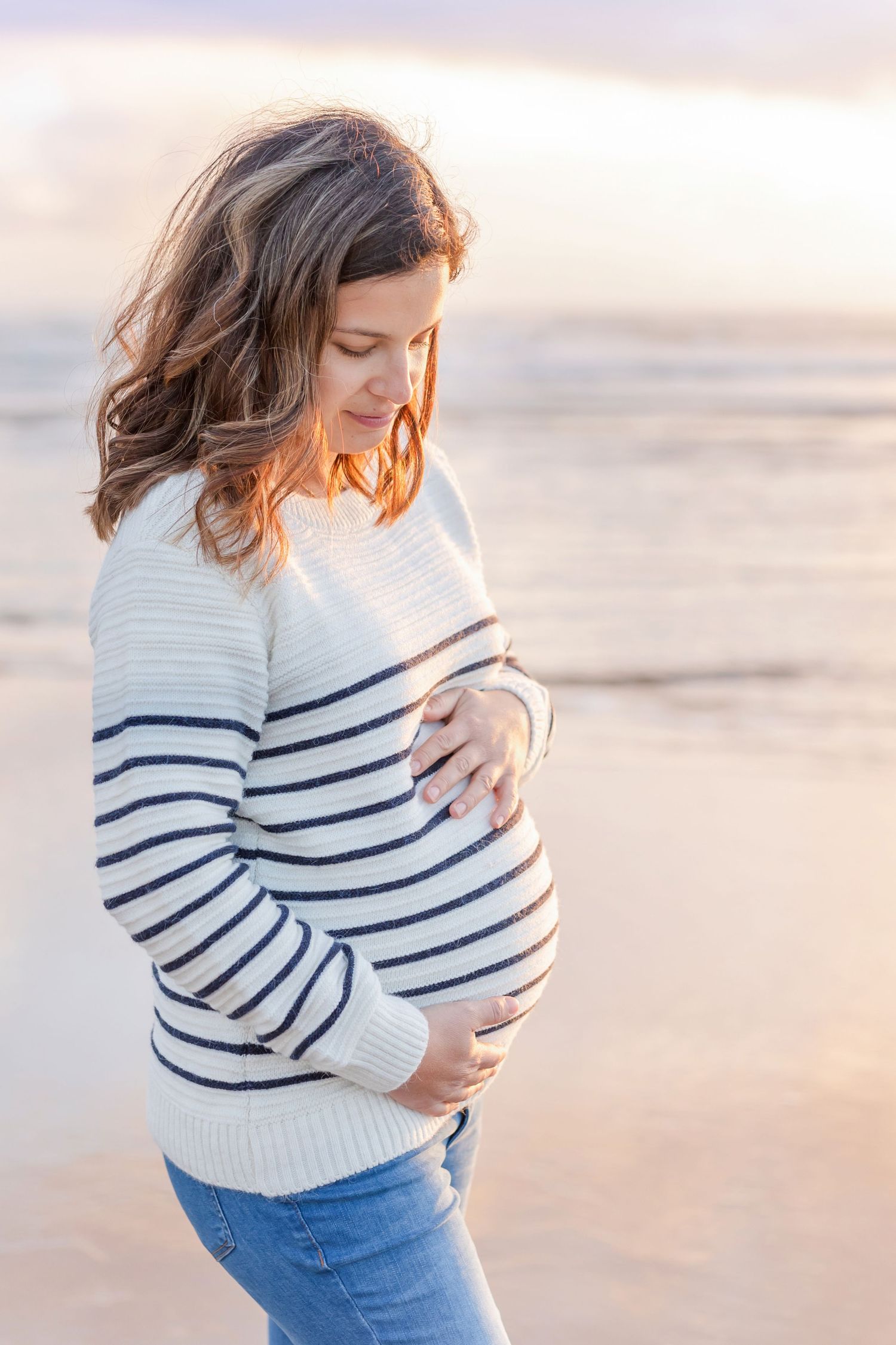 Maternity photo shoot at sunset showing woman in striped sweater caressing baby bump on beach.