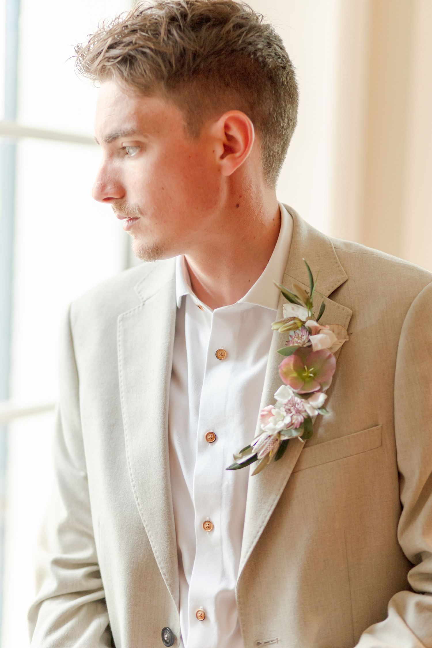 Person wearing a beige suit jacket with floral boutonniere looking towards window.