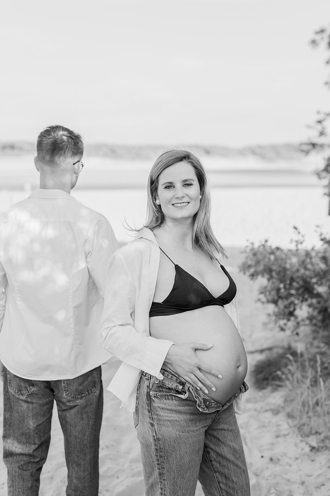 A couple in jeans stands together outdoors by a lake, showing a progression of maternity photos in black and white.