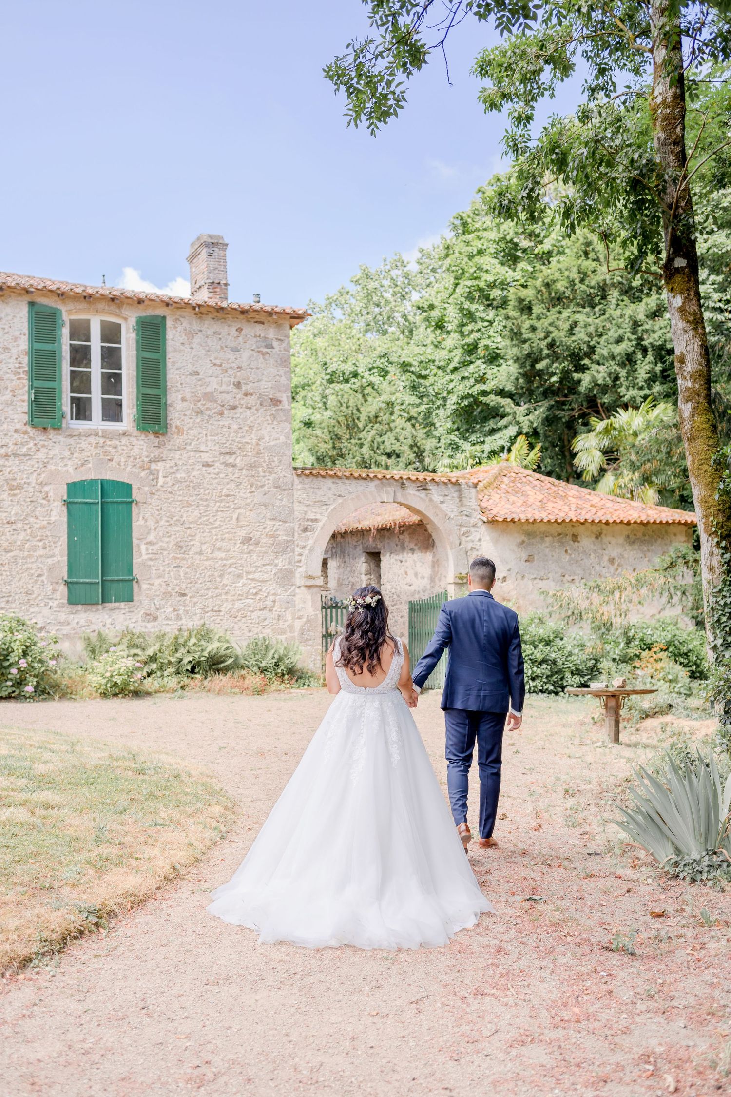 Couple walking along rustic stone path towards Mediterranean-style villa with green shutters.
