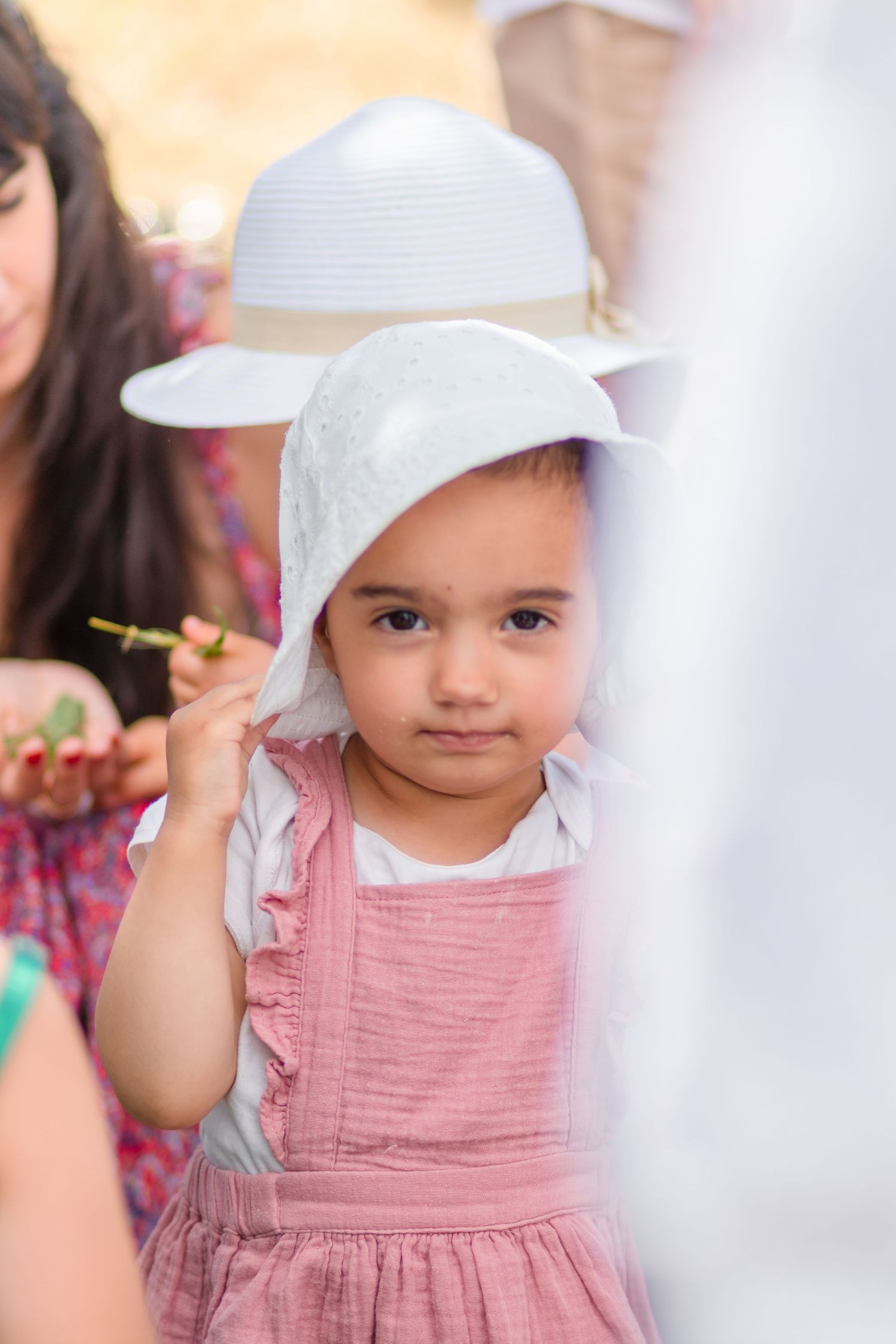 Young child in pink dress and white hat at outdoor gathering.