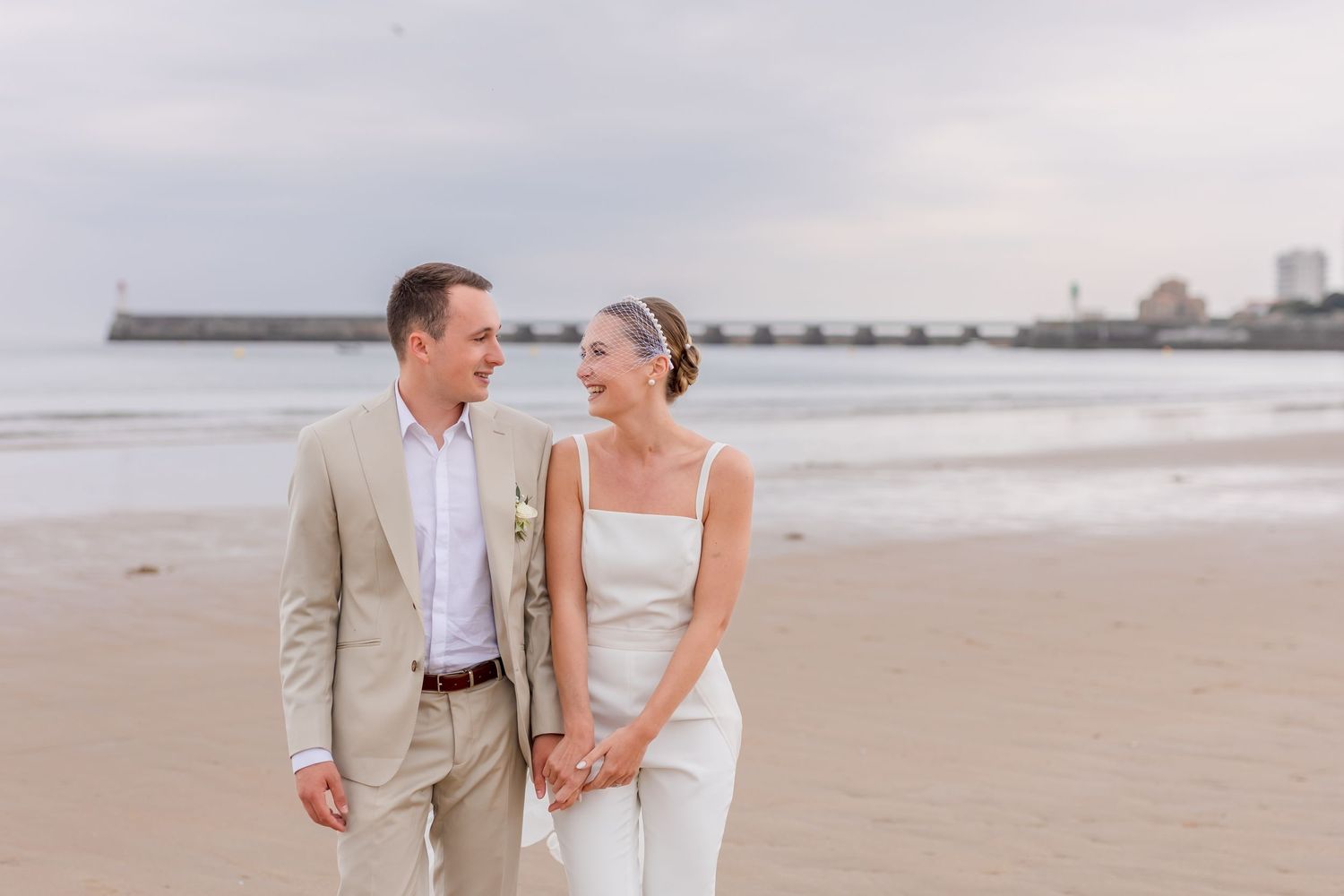 Beach wedding couple walking hand in hand with pier visible in background.