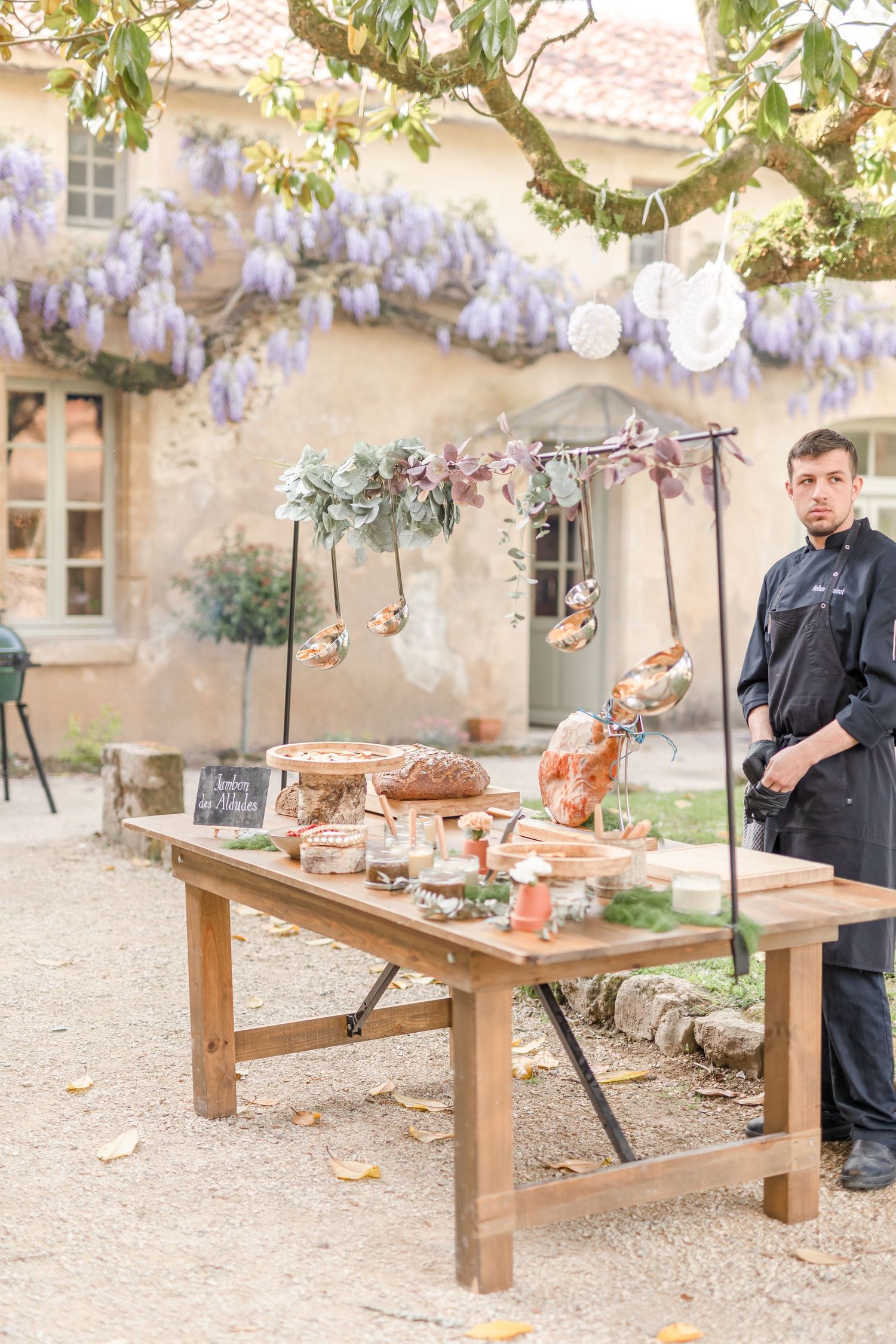 Wooden table setup with catering and drinks under hanging wisteria at an elegant outdoor venue.